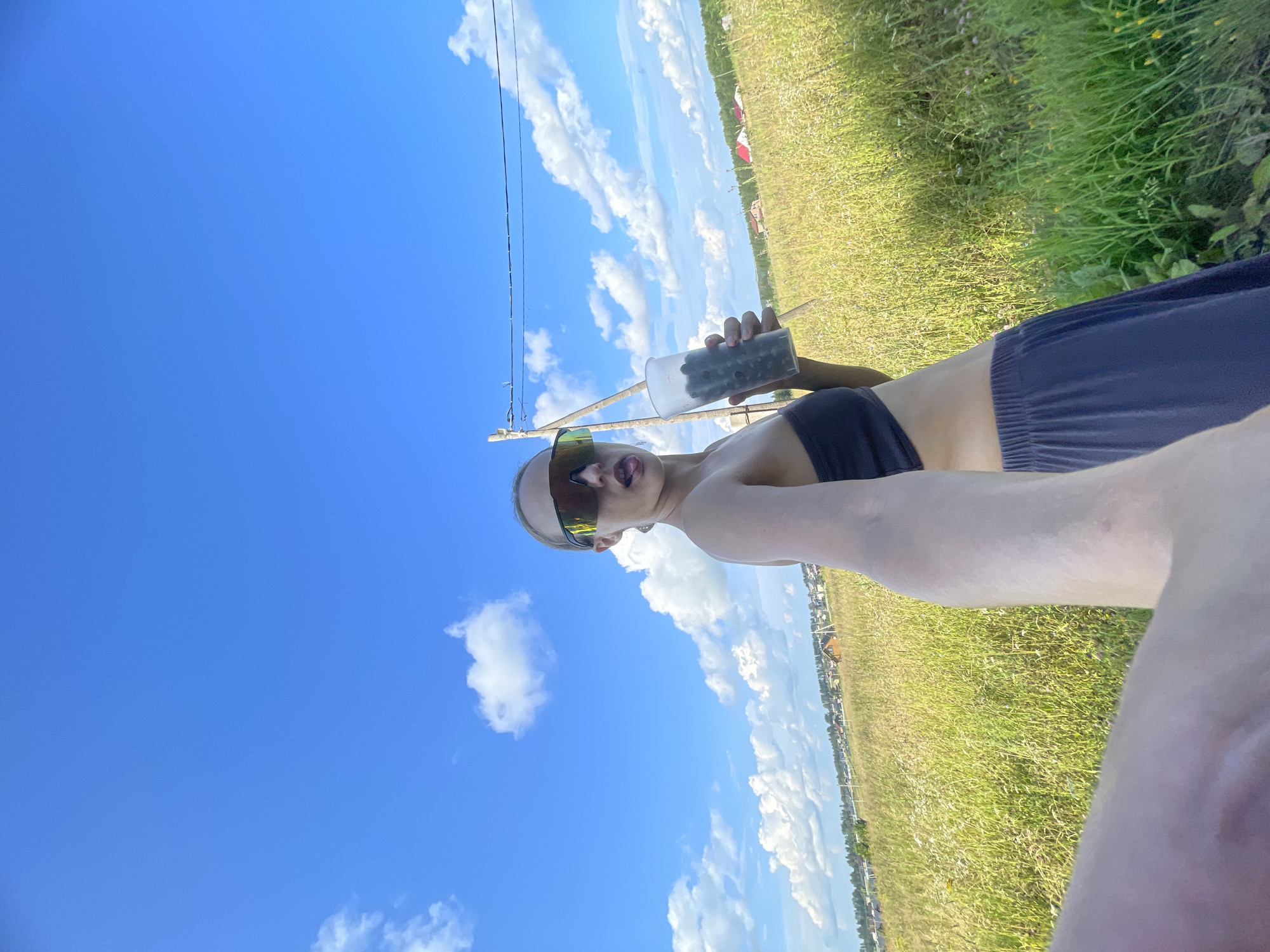 A young Caucasian woman with blonde hair pulled back stands in a sunlit grassy field, holding a clear plastic cup filled with dark berries in her right hand. She is wearing a dark bandeau top and a dark blue, flowy skirt. Her left arm is extended forward, holding the camera, and her face is turned towards it, wearing large, dark-lensed sunglasses. Her tongue is slightly extended from her mouth.
The background depicts a rural landscape with a treeline and houses visible in the distance, suggesting the location is on the outskirts of a settlement. The sky is a bright blue with scattered white, fluffy clouds. A telephone pole with wires runs diagonally across the upper right portion of the frame. The overall scene is consistent with a summer day in a rural area.
Given the context provided, the location is identified as the village of Podshivalovo, Russia.