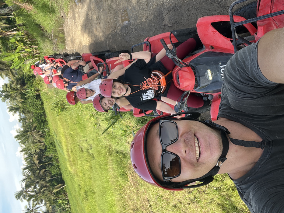 A selfie captures a group of people on red ATVs. The individuals, all wearing red helmets, are lined up and posing for the photo. Lush green vegetation and palm trees fill the background, indicating a tropical location. The person taking the selfie is smiling and wearing sunglasses and a red helmet. The ATVs are red and appear to be Honda brand.