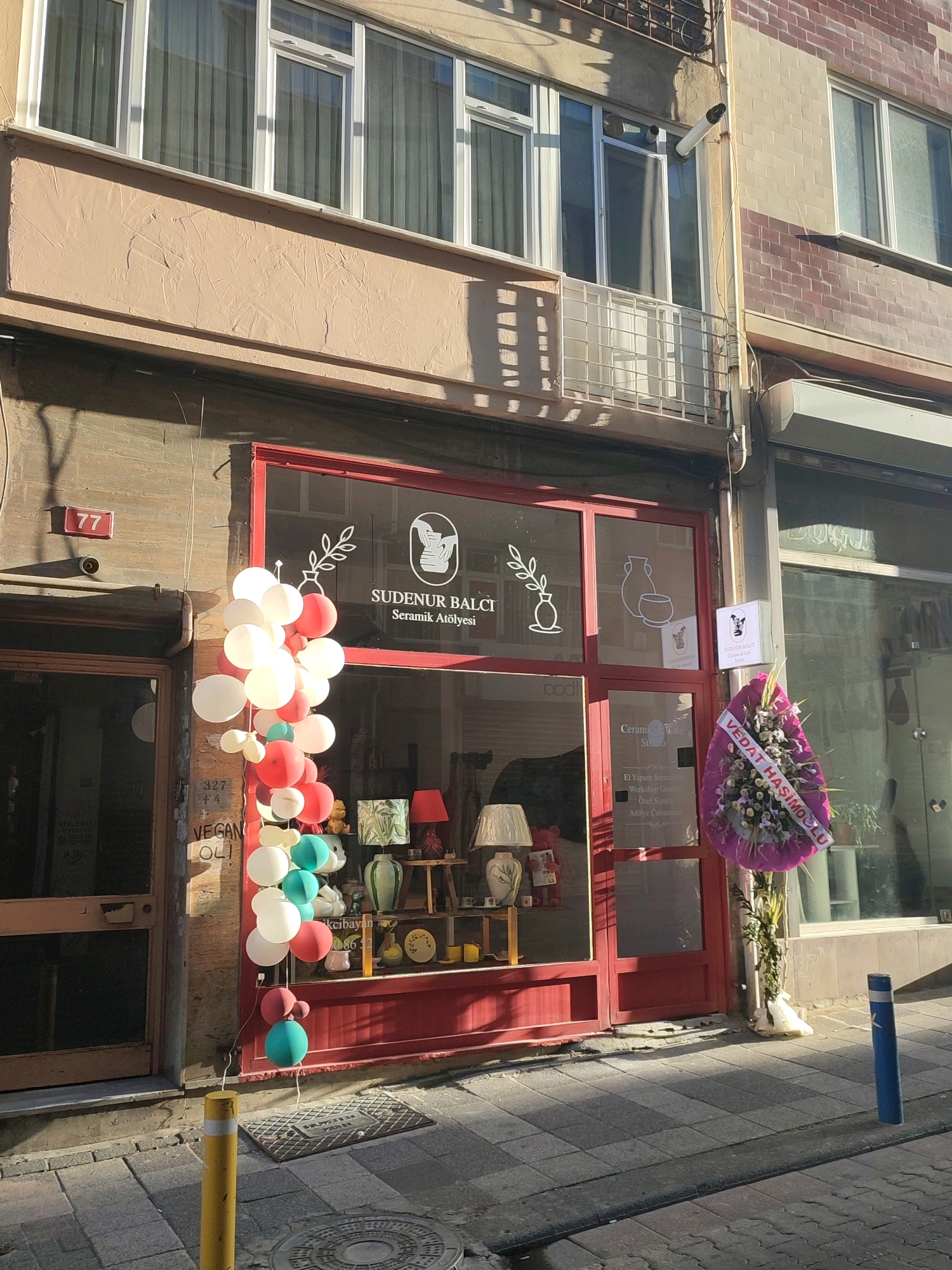 The image shows the exterior of a shop in Kadıköy, Turkey. The shop has a red-framed entrance and window display. A cluster of balloons, predominantly white and red, is positioned to the left of the entrance, extending upwards. A sign above the window reads "SUDENUR BALCI Seramik Atolyesi," indicating it is a ceramic studio. In the window display, there are various decorative items including lamps, ceramic pieces, and stuffed animals. To the right of the entrance, a large, purple wreath with white flowers and a banner that reads "VEDAT HASİMOĞLU" is hanging. The shop is situated on a street with a tiled sidewalk. A yellow bollard is present on the sidewalk in the foreground, and a blue bollard is visible further down the street. The building has a light-colored facade with windows on the upper floors. Sunlight casts shadows on the sidewalk and building.