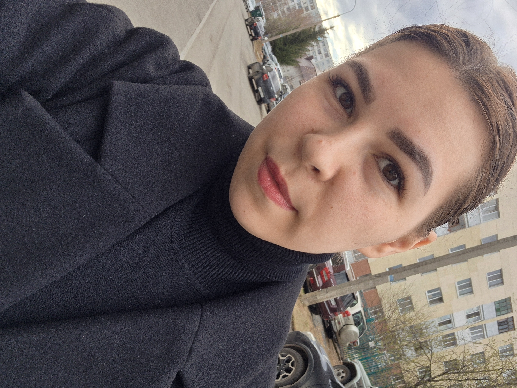 A selfie depicts a young woman in Tobolsk, Russia. She has fair skin, brown eyes, dark eyebrows, and wears a black turtleneck sweater and a black coat. She has light brown hair pulled back. Her lips are tinted a reddish-pink. The background reveals an outdoor urban setting with a row of parked cars on asphalt. A multi-story apartment building with several windows and beige and brown siding is visible. Bare tree branches are in the foreground. The sky is overcast.
