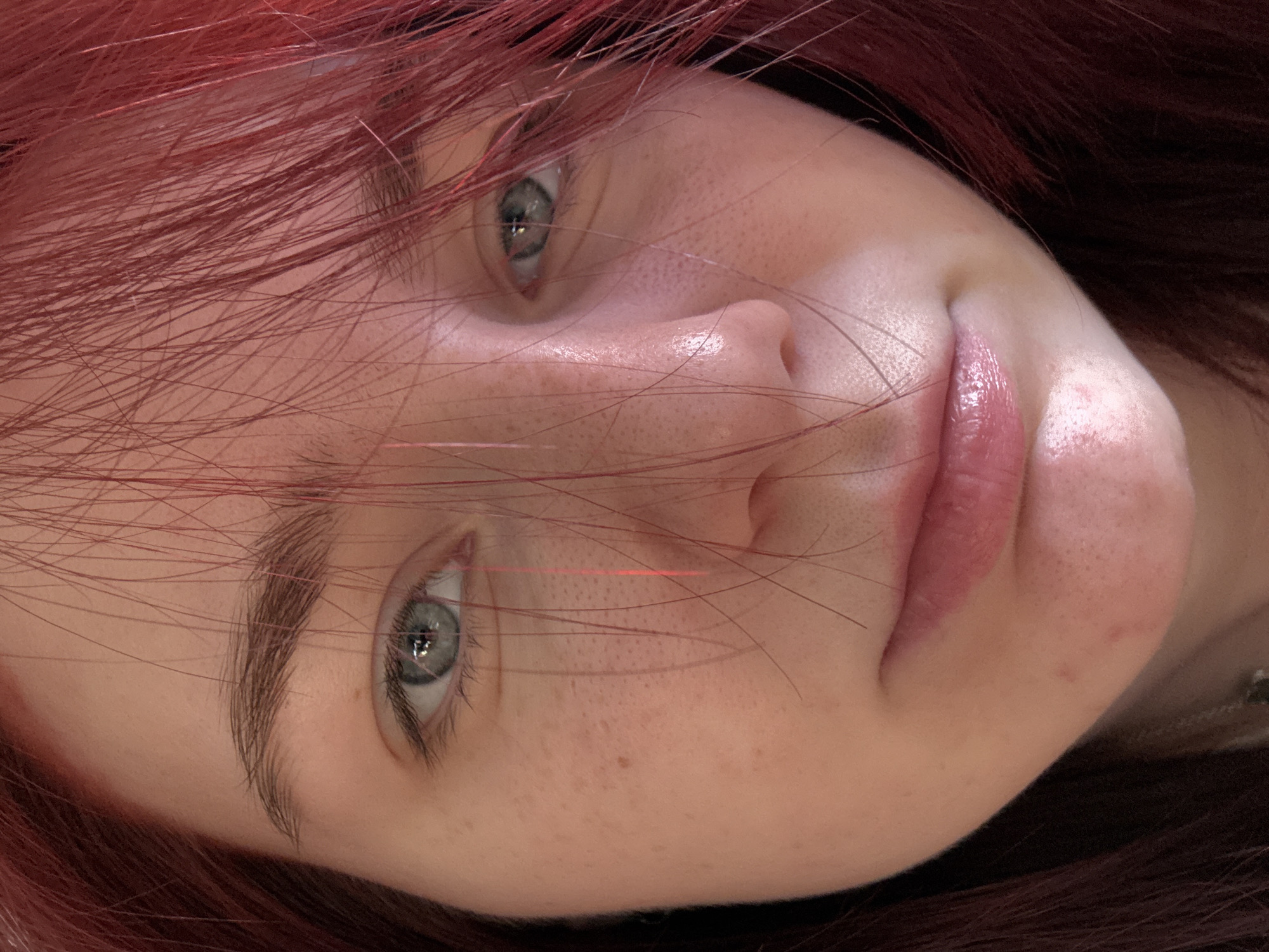 A close-up, indoor, head-and-shoulders shot features a young woman with reddish-brown hair that is partially obscuring her face. Her skin is fair with a smattering of freckles across her cheeks and nose. Her eyes are a light blue-gray, with visible eyelashes and eyebrows. Her lips are slightly parted and appear to be glossed. The background is indistinct, suggesting an indoor setting with soft lighting.