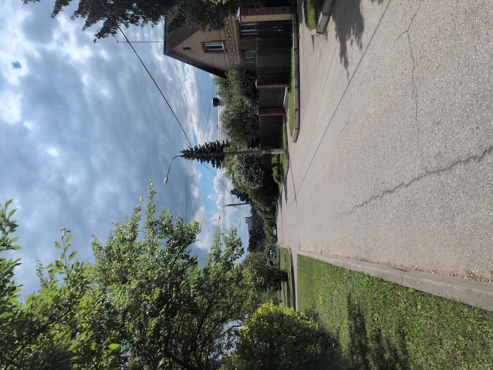 The image displays a quiet residential street in Dubrovitsy, Russia, under a partially cloudy sky. The perspective looks down a paved asphalt road that extends into the distance on the right side of the frame. The road surface is light gray, showing some cracks. It is bordered on the right by a gray concrete curb and a well-maintained green grassy verge, with tree shadows stretching across both.
On the immediate right, a two-story house is prominent, constructed from light brown bricks with a gabled brown roof and rectangular windows. A dark wooden fence marks the property line, partially hidden by dense green foliage.
The left side of the frame features abundant green trees, including broad-leaved deciduous types and a tall, slender evergreen tree, possibly a fir or spruce. A modern street light is affixed to a black pole on this side of the road, and multiple black utility wires crisscross against the sky. The sky above is a mix of bright blue patches and white to gray clouds. In the far distance, faint outlines of additional buildings suggest a wider settlement.
The scene depicts a peaceful, typical daytime setting. There are no people or specific activities taking place. The weather appears fair and bright despite the cloud cover. No visible text is present in the image.