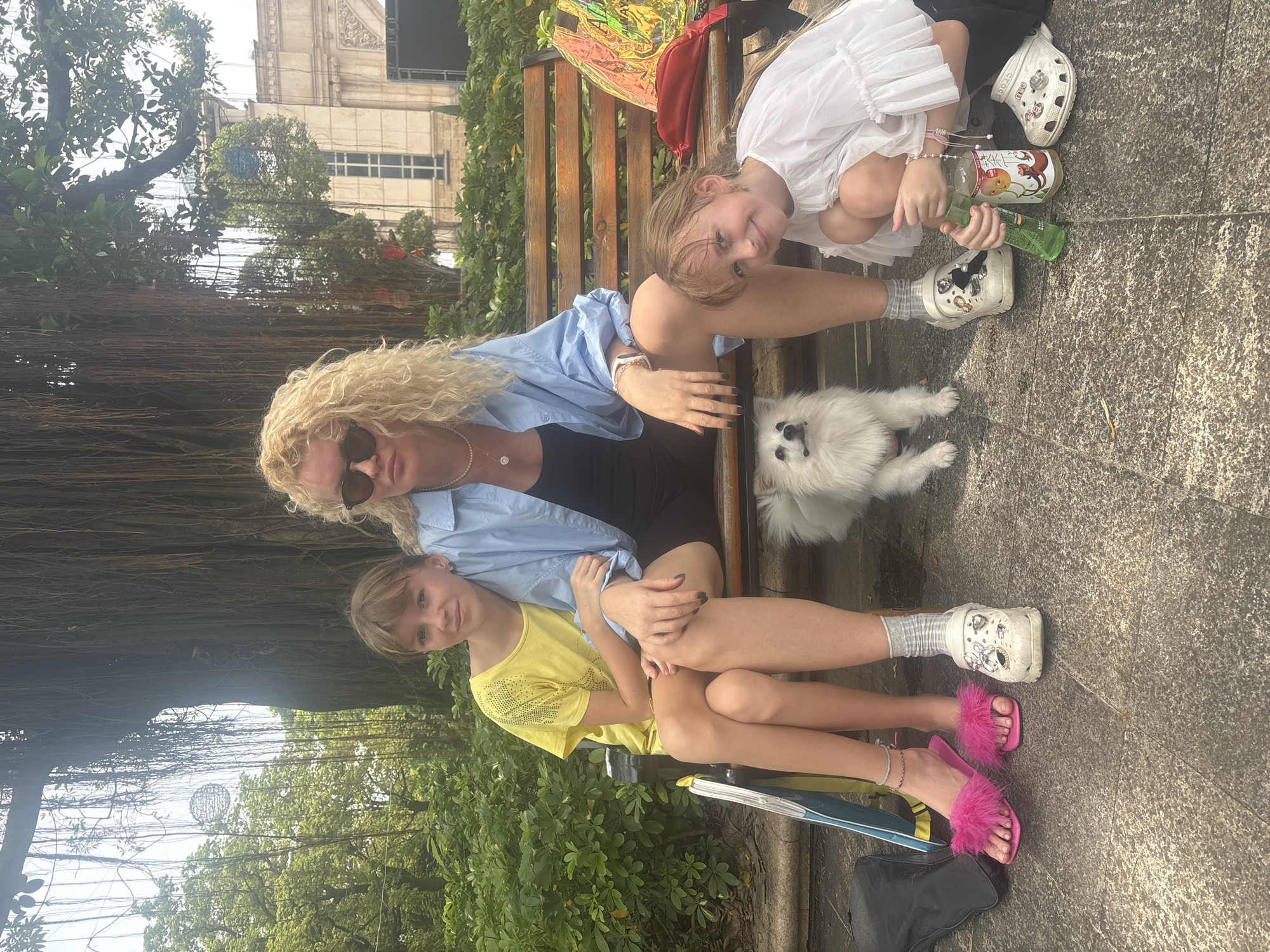 The image depicts three people and a dog posing for a photograph on a wooden bench in an outdoor setting, possibly a park or public garden in Nanhai District, China. A woman with curly blonde hair and sunglasses is seated in the center, wearing a light blue open shirt over a black top and dark shorts. To her left, a young girl with light brown hair and a yellow t-shirt is seated, looking towards the camera. To the woman's right, another young girl with light brown hair is kneeling on the ground, wearing a white top with ruffled sleeves and black shorts. She is holding two bottles, one green and one with a label featuring cartoon characters. A small white dog sits on the ground between the woman and the girl on the left. The background features lush green foliage and the hanging roots of a large tree, with hints of a building in the distance. The ground is paved with textured stone. The overall lighting suggests it is daytime, possibly overcast.