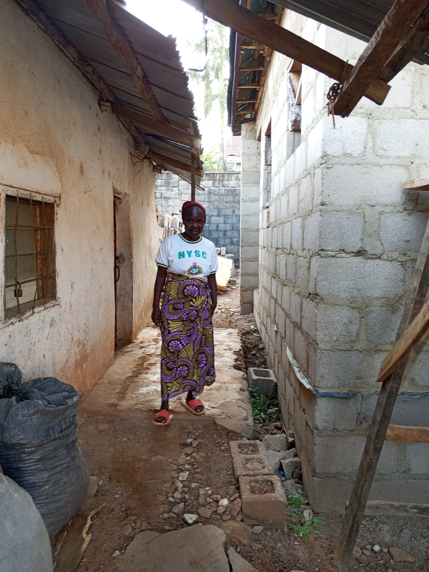 A woman stands in a narrow passageway between two buildings. She is wearing a white t-shirt with "NYSC" printed on it, a colorful, patterned skirt, and red sandals. She has a red head covering. The building on the left is weathered and appears older, with a window and a doorway visible. Several large, dark bags are near the building on the left. The building on the right is constructed of cinder blocks and is unfinished, with some wooden planks nearby. The passageway is unpaved and littered with debris. The scene is in Bukuru, Nigeria.