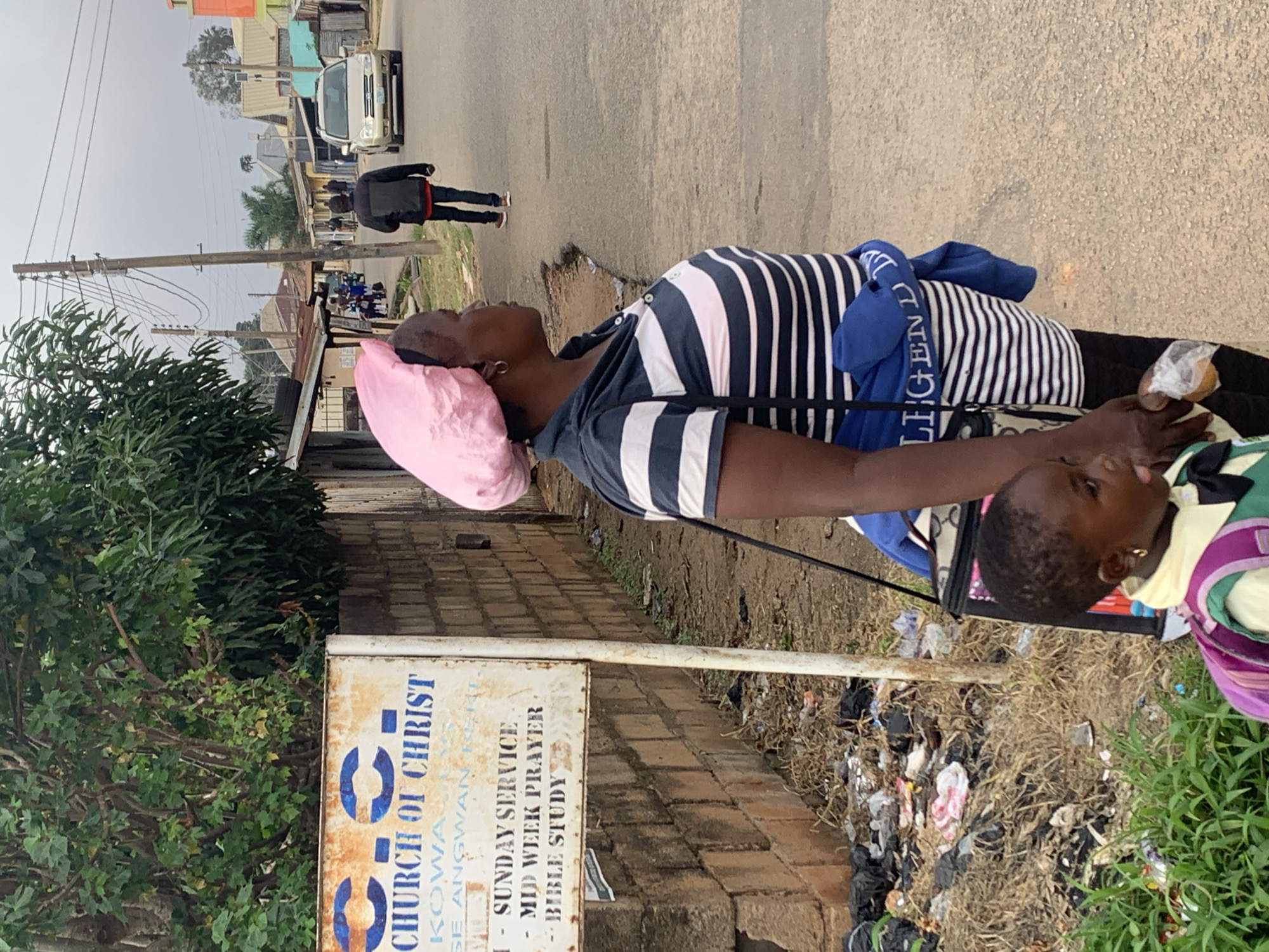 A woman in a pink headscarf and a striped shirt stands with a child in the foreground, facing away from the camera towards a street in Jos, Nigeria. A sign for the Church of Christ is visible to the left of the woman, and a pile of trash is on the ground next to it. In the background, cars and people are walking along the street, with buildings lining either side. The overall scene depicts an urban environment with signs of neglect and everyday life.