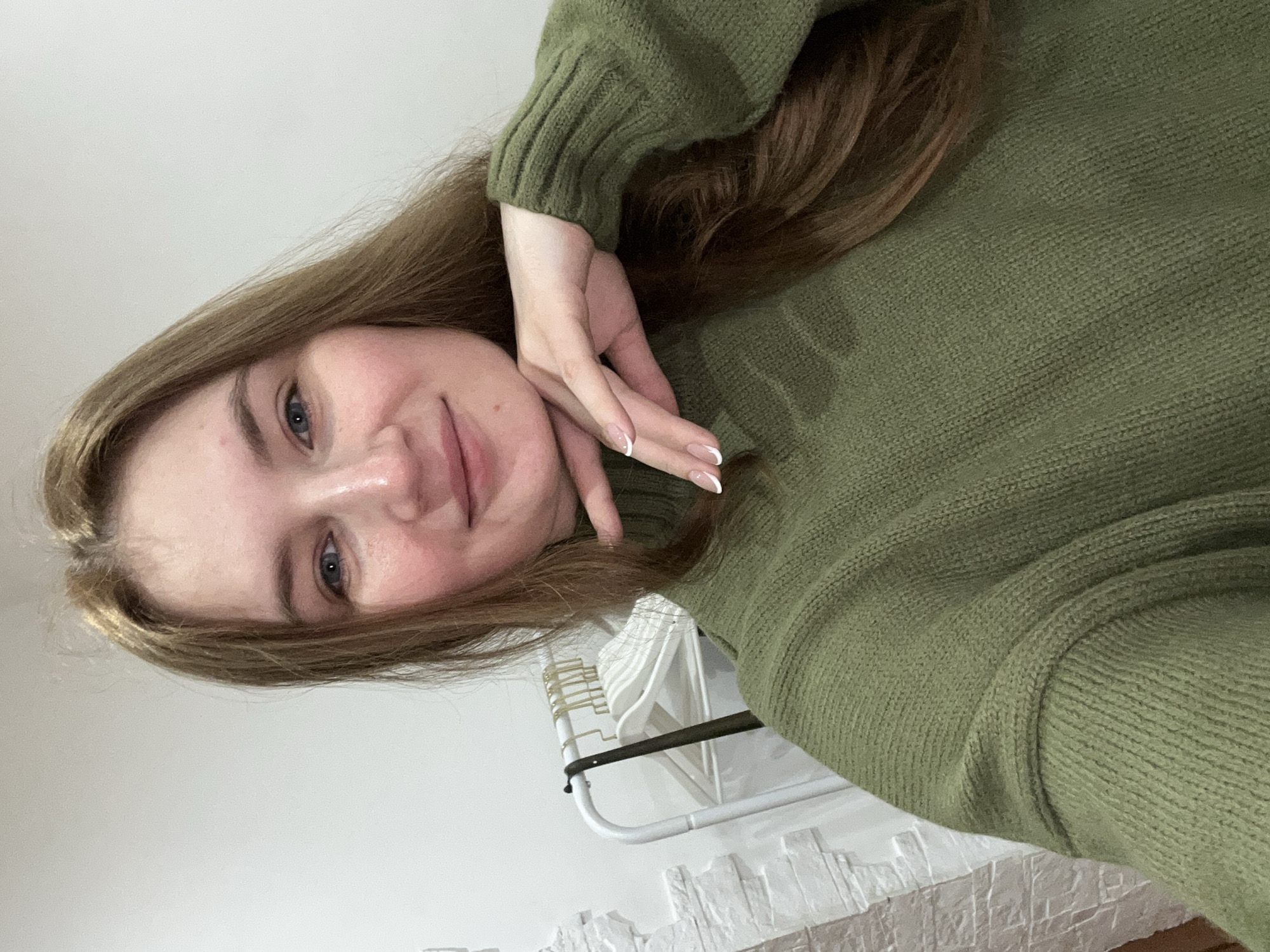 A young woman is featured in an indoor selfie, looking directly at the camera with a gentle, understated smile. Her head is tilted, giving the image a casual and relaxed feel. She has long, light brown hair and light-colored eyes, appearing blue or grey. She is dressed in a cozy, olive green knitted turtleneck sweater, with the texture of the fabric clearly visible. Her right hand is raised towards her face, resting near her chin and neck, showcasing neatly manicured nails with a distinct white French tip design.
The background is minimalist, consisting mainly of a plain white wall. To the right of the frame, a white clothes rack with several hangers is partially visible. Below the clothes rack, a textured white surface, resembling decorative brickwork or a radiator cover, adds a subtle detail to the lower right portion of the image. The lighting is soft and even, suggesting a well-lit interior space. The overall impression is one of calm and comfort. The image is rotated 90 degrees clockwise. No text is visible in the frame. The setting is an indoor location in Chaikovsky, Russia.