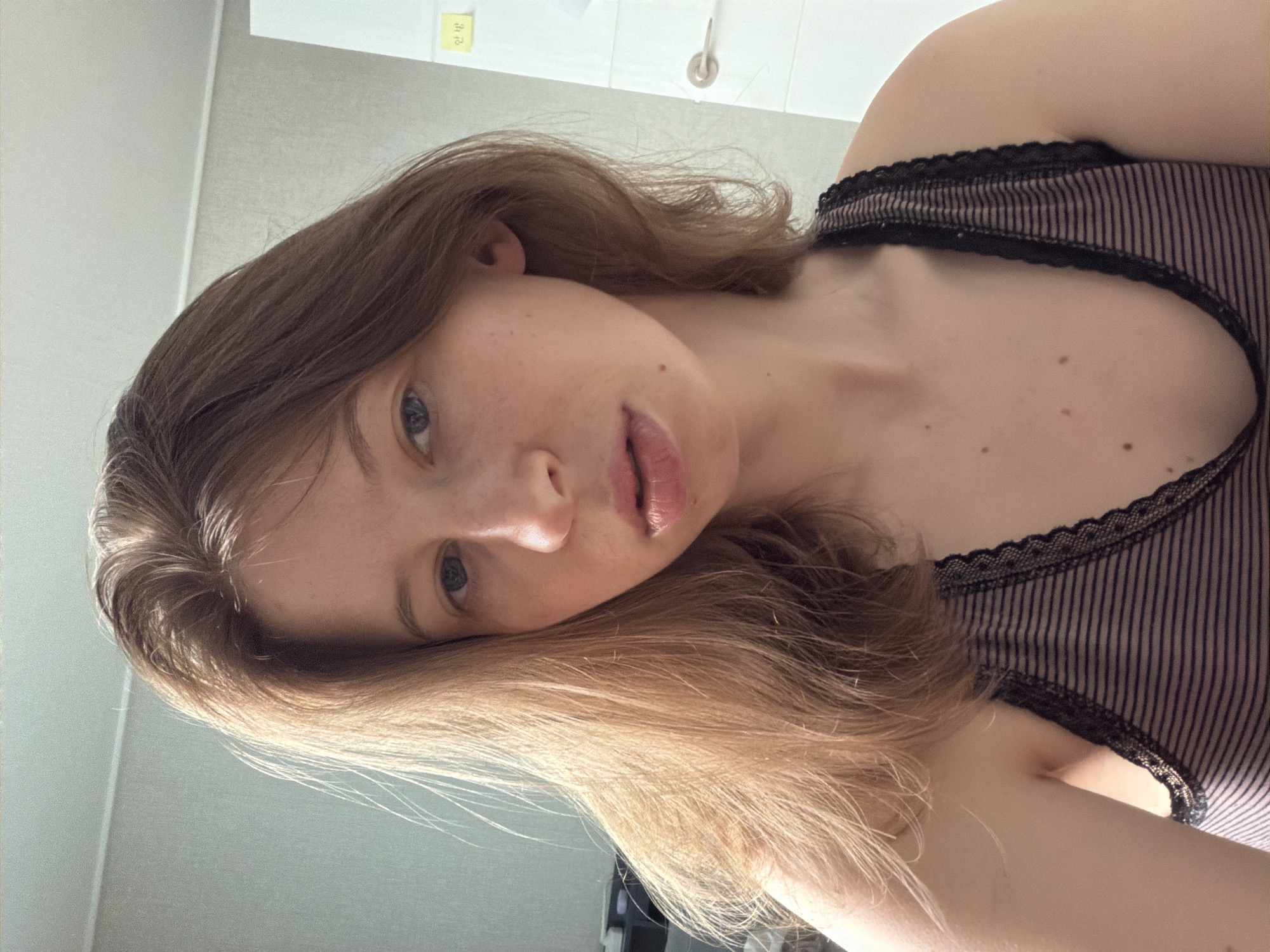 The image is a close-up, self-taken photograph of a young Caucasian woman with light brown hair. She is wearing a striped tank top with lace trim. Her expression is neutral, and she is looking directly at the camera. The background appears to be an indoor setting, possibly a room in an apartment or house, with a wall and a door visible. A small yellow sticky note with Korean writing is attached to the door. The lighting suggests it is daytime. The image was taken in Dongducheon-si, South Korea.