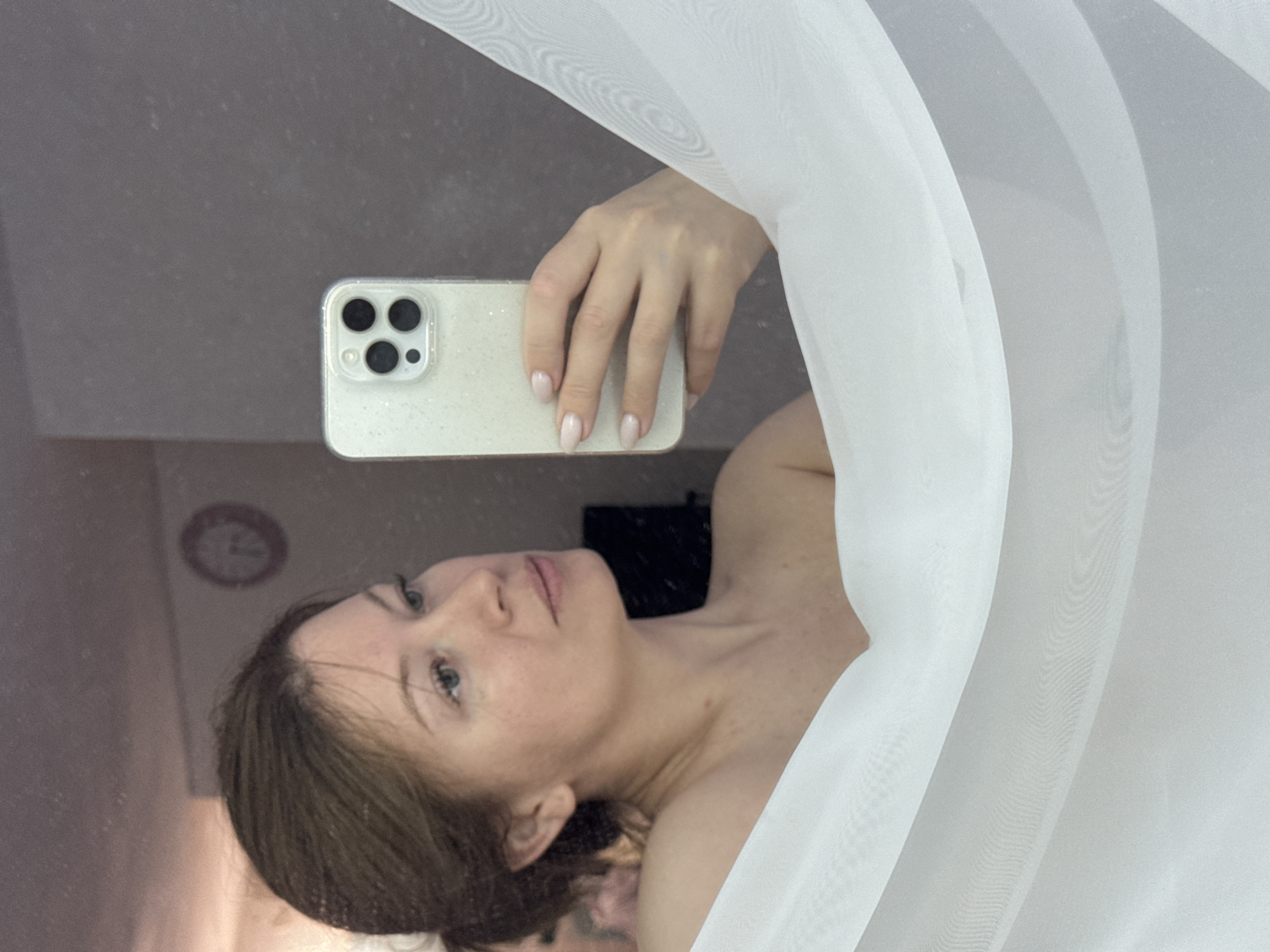 A woman is taking a selfie in a mirror. She is holding a white iPhone in her right hand. A white sheer curtain is draped around her body. A clock is visible on the wall in the background. The location is likely Chaikovsky, Russia.