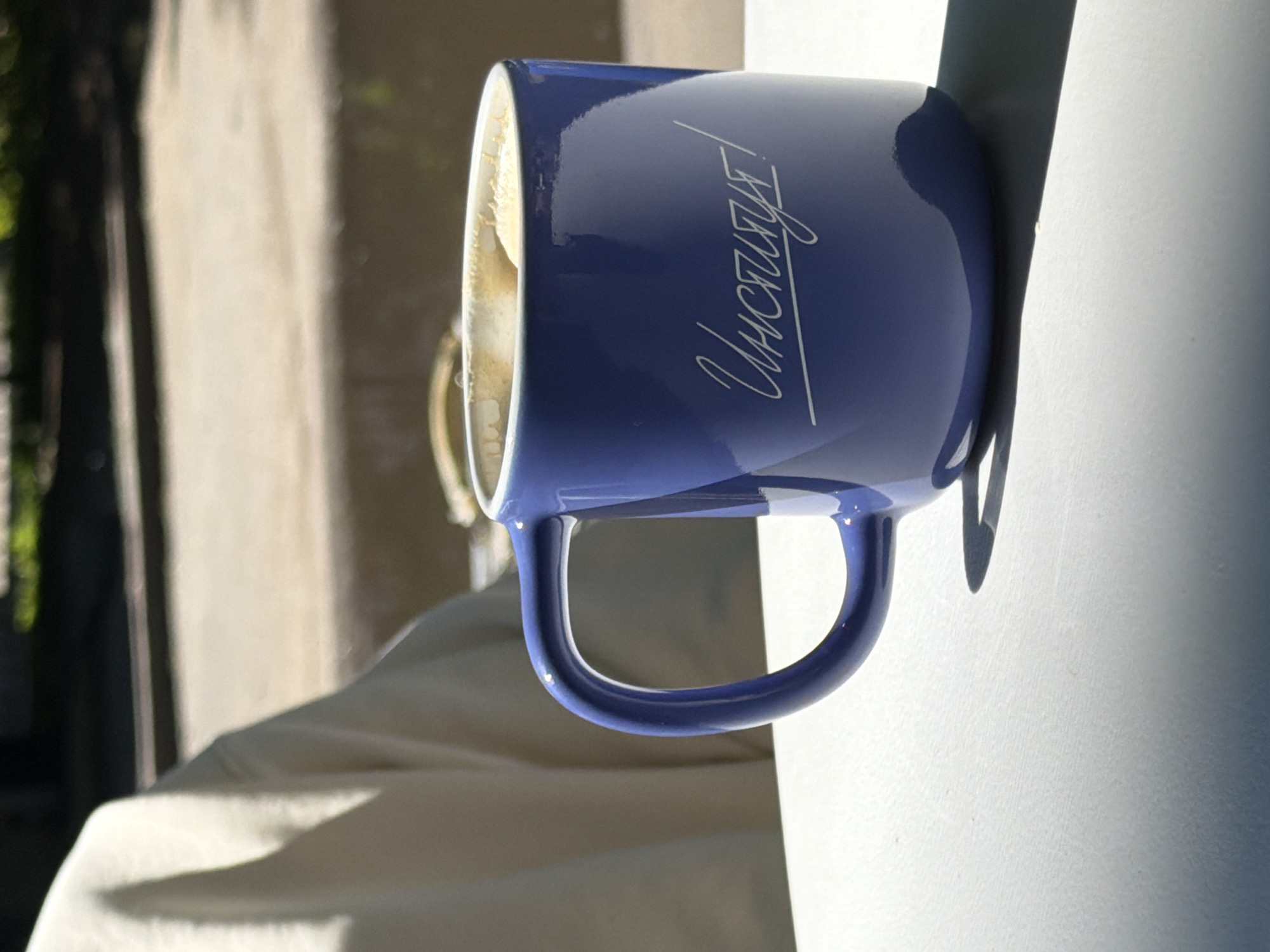 A close-up, eye-level shot shows a blue ceramic mug with white lettering that reads "Институт!" (Institute!). The mug is filled with a light-brown beverage, possibly coffee, with a frothy top. The mug is positioned on a white surface, casting a shadow to its right. In the background, a person's shoulder and upper arm are visible, wearing a cream-colored garment. The environment appears to be outdoors, with a concrete wall and greenery in the blurred background, suggesting a public space in Saint Petersburg, Russia, possibly near a park or an educational institution. The lighting indicates it is daytime.