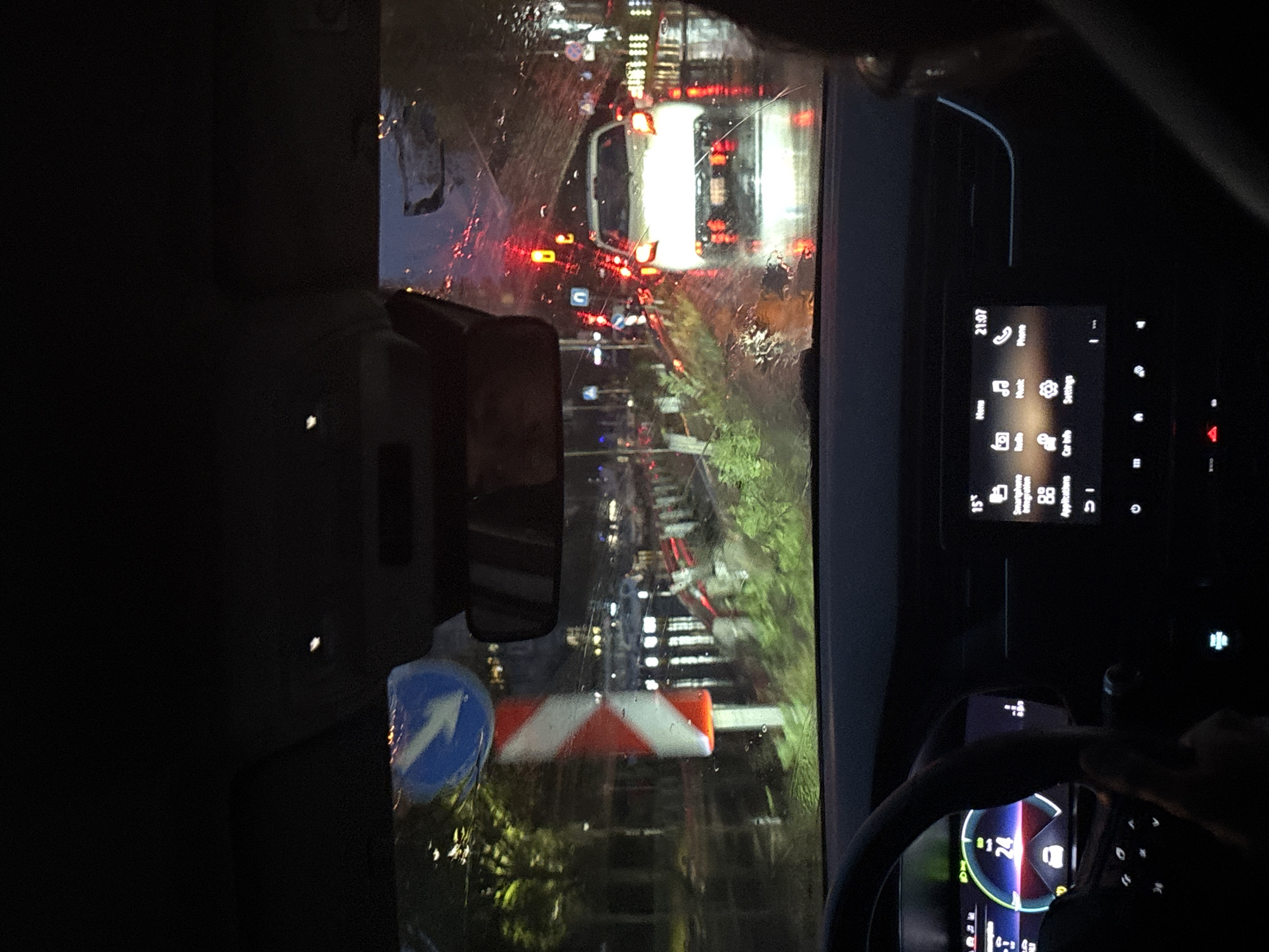 The image, taken from inside a car at night, depicts a view through a rain-streaked windshield onto a busy road. The perspective is that of a driver or front-seat passenger.
Inside the vehicle, the dark interior shows a car's modern infotainment screen on the right, displaying the time "21:07". Menu options like "Phone", "Settings", "Car Info", "Smartphone Integration", and "Applications" are visible, along with a temperature of "15°". Below the screen, a hand is partially seen gripping the steering wheel, and the digital instrument cluster shows a speed of "24" (likely km/h or mph). A rearview mirror is partially visible on the left.
Outside, the wet windshield distorts the view of the traffic ahead, giving the lights a diffused and streaky appearance. Several vehicles are caught in traffic, indicated by their bright red taillights and headlights. A prominent white vehicle, possibly a van or truck, is directly in front. On the side of the road, a blue circular mandatory sign with a white arrow pointing straight or right is visible. Further along, a red and white chevron barrier or sign is present. The urban setting is suggested by the numerous artificial lights from buildings and streetlights, blurred by the rain.
The activity is driving or being a passenger in a car during nighttime traffic. The weather is rainy, causing reflections and distortions on the windshield. No specific location can be definitively identified, but it appears to be an urban environment. The overall mood is typical of night driving in wet conditions; there are no strong emotional cues.