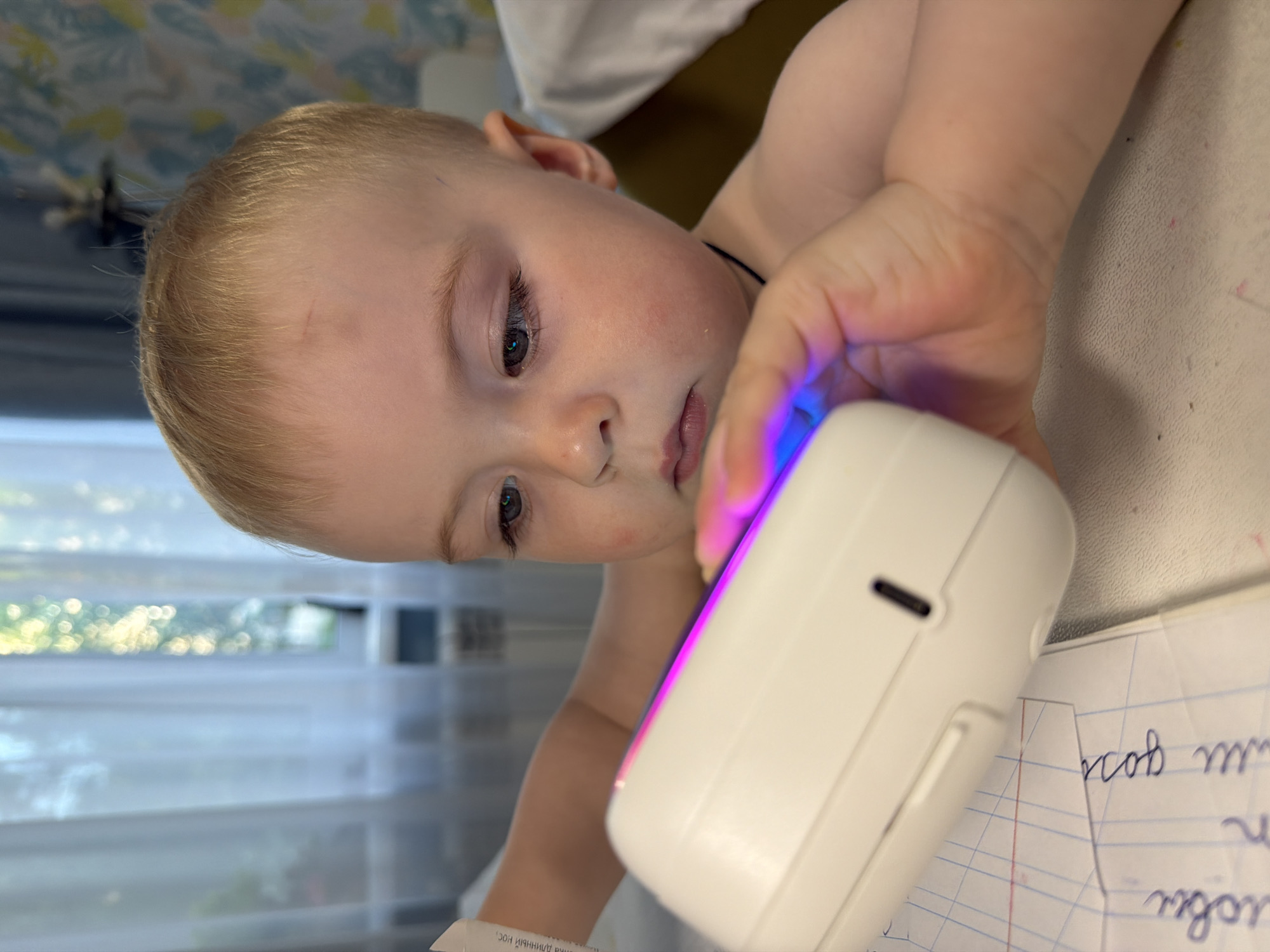 A child lies prone on a surface near a window, interacting with a white, rectangular electronic device emitting a blue and pink light. The child has fair skin, blonde hair, and appears to be looking at the device. A piece of paper with writing in Cyrillic script is visible under the device. The location is possibly in Votkinsk, Russia, though this is not directly discernible from the image alone.