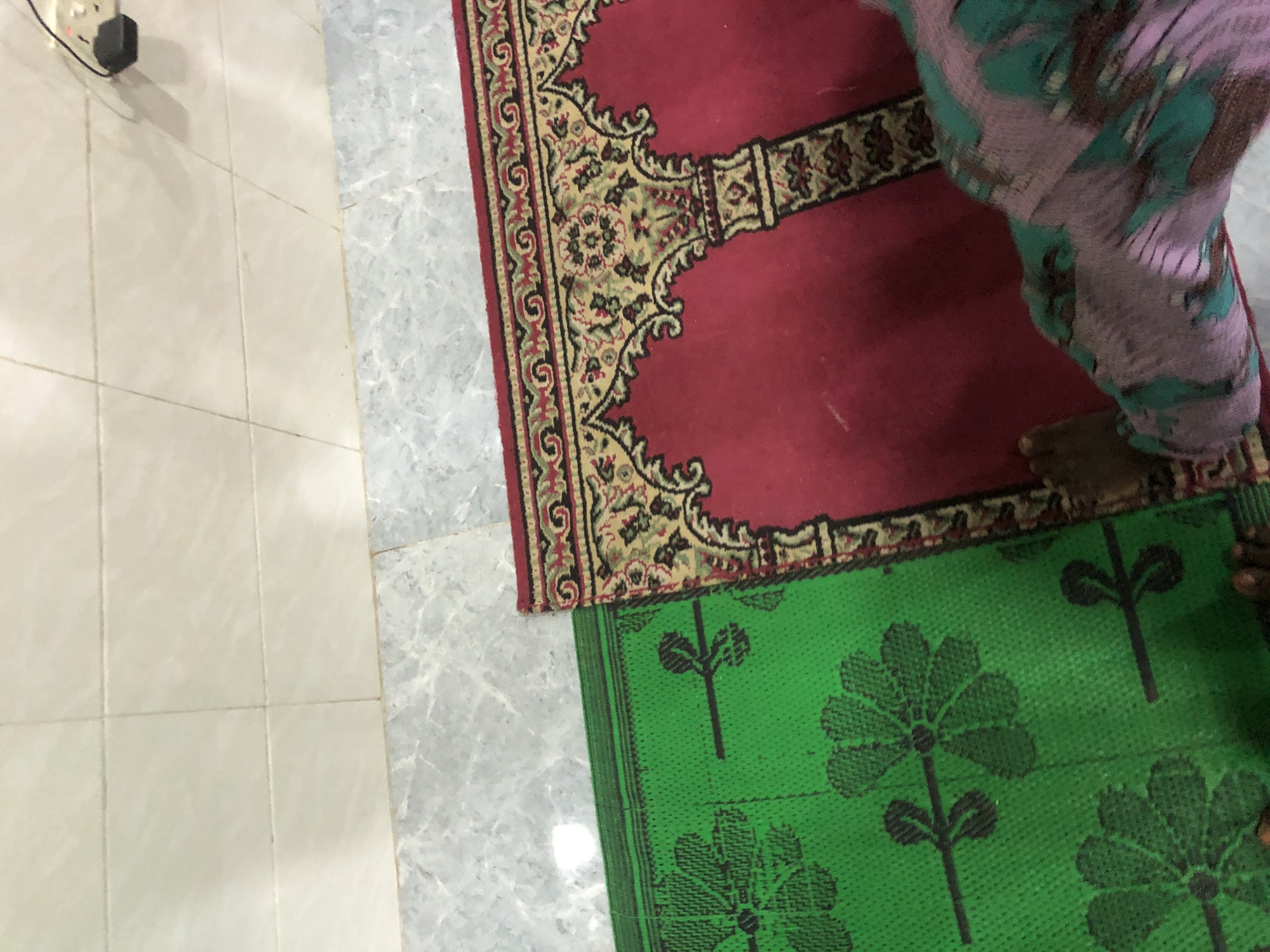 The image shows two prayer mats on a tiled floor. The mat on the right is red with an ornate gold and black pattern featuring arches. The mat on the left is green with a repeating floral pattern in black. A person's feet and lower legs are visible on the right side of the image, standing on the red prayer mat. The person is wearing a patterned garment. The flooring appears to be a combination of light beige tiles and marble-patterned tiles. In the upper right corner, a white electrical outlet with a black plug is visible on the beige tiles. The scene appears to be indoors. The provided location is Ado, Nigeria.