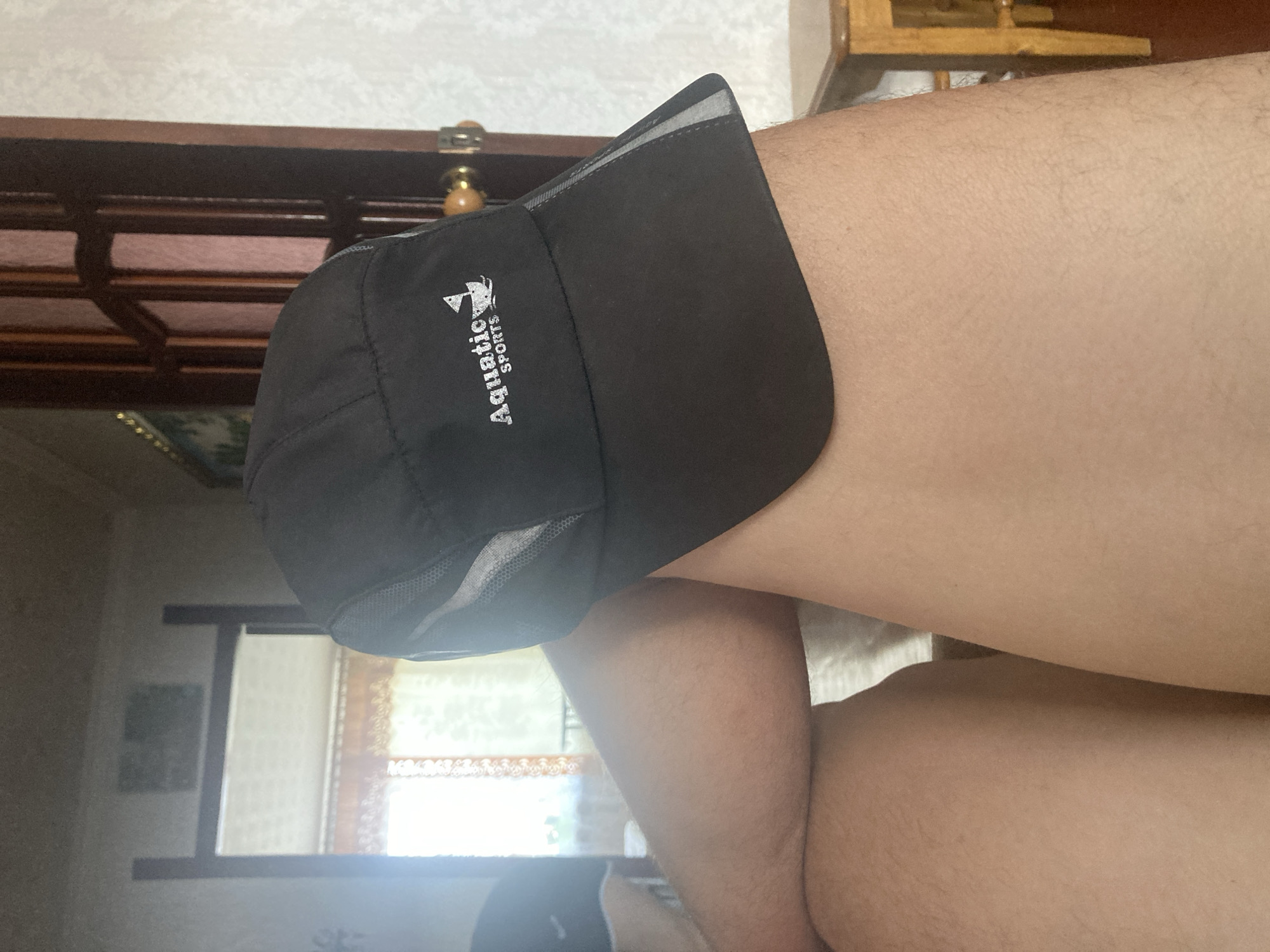 A black baseball cap with the text "Aquatic SPORTS" and a small sailboat graphic is placed on top of a person's thighs. The cap is positioned diagonally, with the brim facing towards the viewer's left. The background shows a room interior with a window, curtains, and a wooden door. To the right, a small wooden table is partially visible. The lighting suggests it is daytime, with light coming from the window. There are no other people or animals visible in the image. The scene is likely indoors, possibly in a residential setting.
