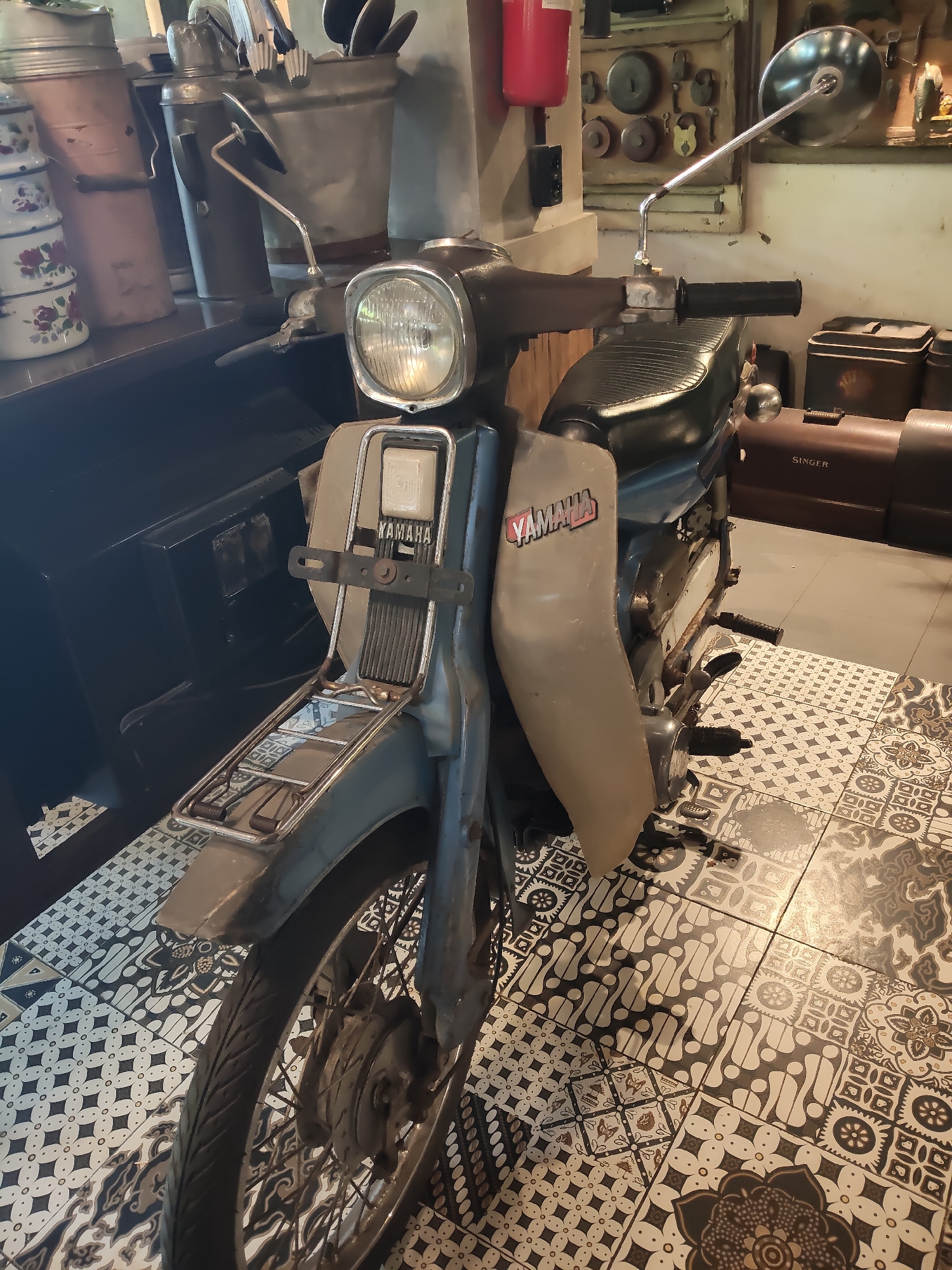 A vintage Yamaha motorcycle, predominantly blue and grey, is parked indoors on a floor tiled with an intricate, patterned design featuring dark brown, white, and light beige elements. The motorcycle faces forward and slightly to the left. It has a round headlight, a black seat, and a Yamaha logo on its side panel. Behind the motorcycle, the environment appears to be a shop or display area. On a shelf to the left, there are stacks of ceramic containers with floral patterns and a tall metal canister. To the right, several vintage metal boxes, including one with a "Singer" logo, and a wall display of old locks and keys are visible. The lighting suggests an indoor setting with some natural light entering from the right. There are no people or animals present in the image. The scene is static and appears to be a curated display of vintage items.