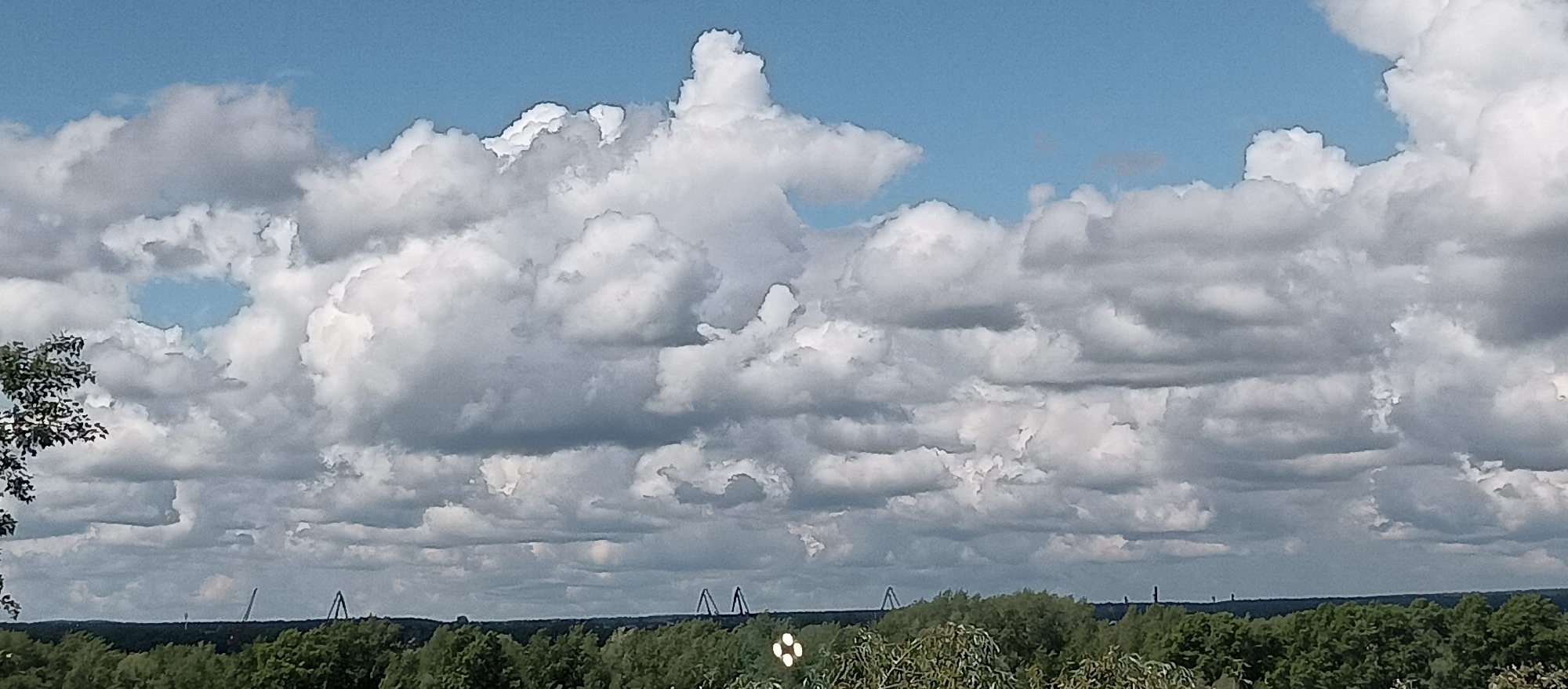 The image depicts a landscape under a partly cloudy sky. The sky is mostly blue, with a significant portion covered by white and grey clouds. In the foreground, a line of green trees extends across the horizon. Above the trees, several industrial structures or towers are faintly visible. The overall environment appears to be outdoors, likely a natural or park-like setting within the city of Unknown, Unknown.