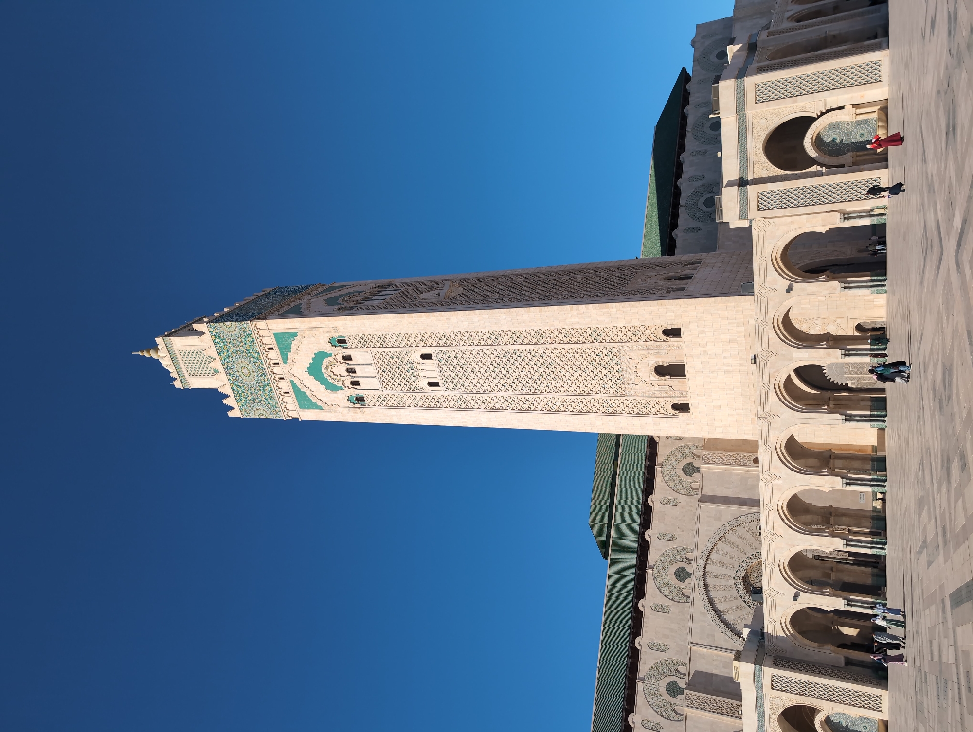 The image shows the Hassan II Mosque in Casablanca, Morocco. The mosque is a large, intricately designed building with a tall minaret. The minaret is decorated with green and white tiles, and the mosque itself is made of beige stone. Several people are walking around the mosque. The sky is clear and blue.