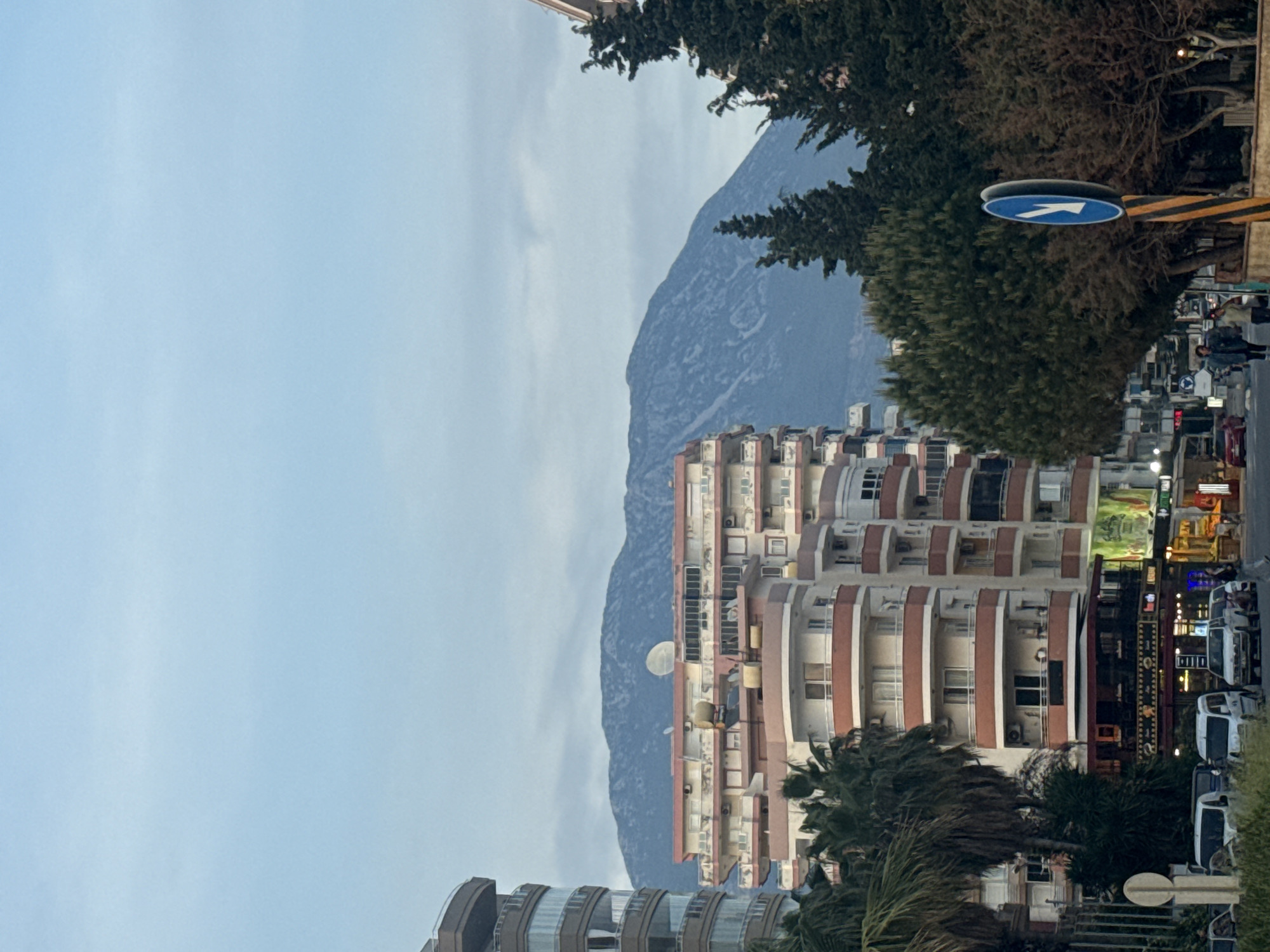 The image shows a multi-story building with balconies, likely a residential or commercial structure, situated in an urban environment with a mountain backdrop. A street sign is visible on the right, along with signs and traffic lights, and vehicles parked along the road. Trees and other vegetation are present near the buildings and street. The sky is overcast. No people are prominent, but there may be some visible in the background along the street. The location is unknown.