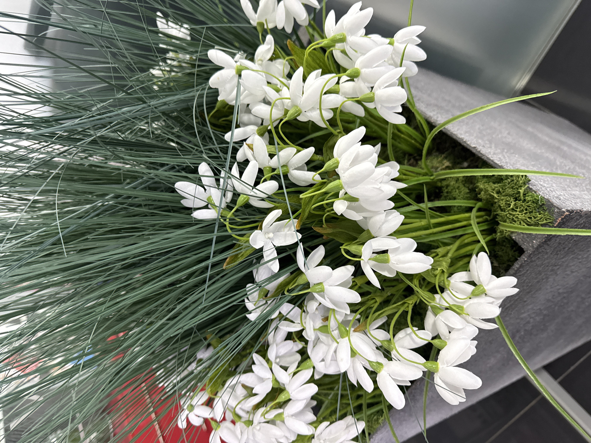 The image shows a close-up, high-angle shot of an arrangement of artificial snowdrop flowers and green grass-like foliage, likely displayed indoors. The snowdrops are white with closed petals. The arrangement is in a gray container. The photograph's location cannot be identified as Perm, Russia, from the visual content alone.
