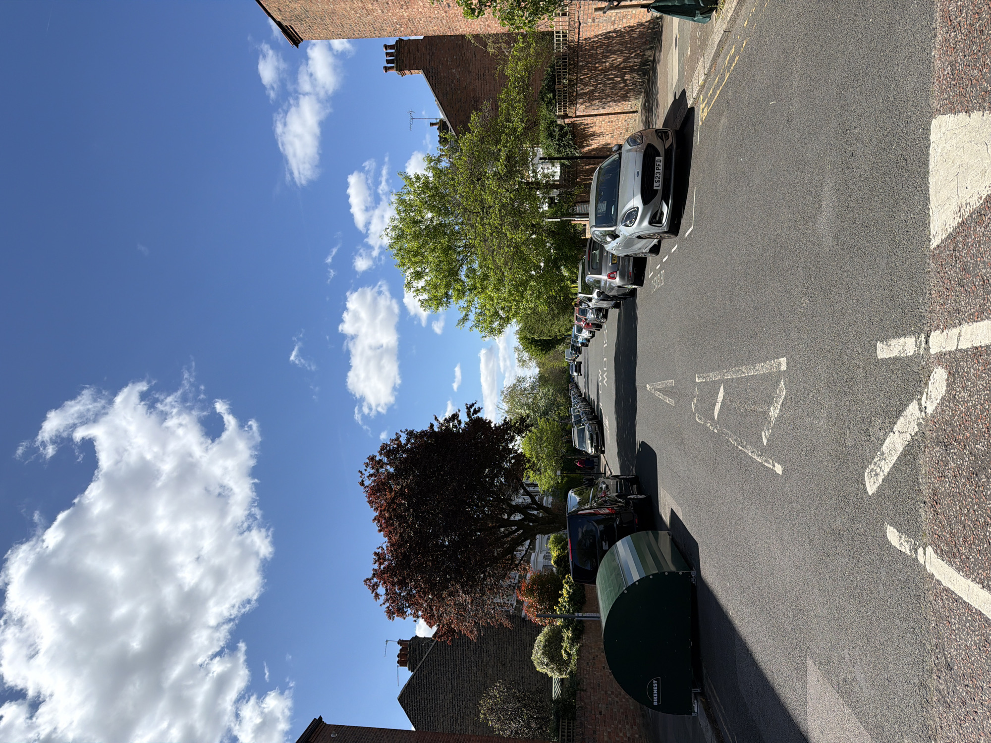 The image depicts a residential street in London, United Kingdom, on a sunny day with scattered clouds in a bright blue sky. The road is lined with parked cars on both sides. A silver Ford Fiesta with the license plate LS23 PFD is parked closest to the viewer on the right side of the street. The road has road markings, including painted white triangles and a broken white line down the center. On the right, a green "BikeNest" bicycle storage unit stands next to the sidewalk. Brick buildings are visible on either side of the street, interspersed with trees, one of which has maroon leaves. Other cars are parked further down the street.