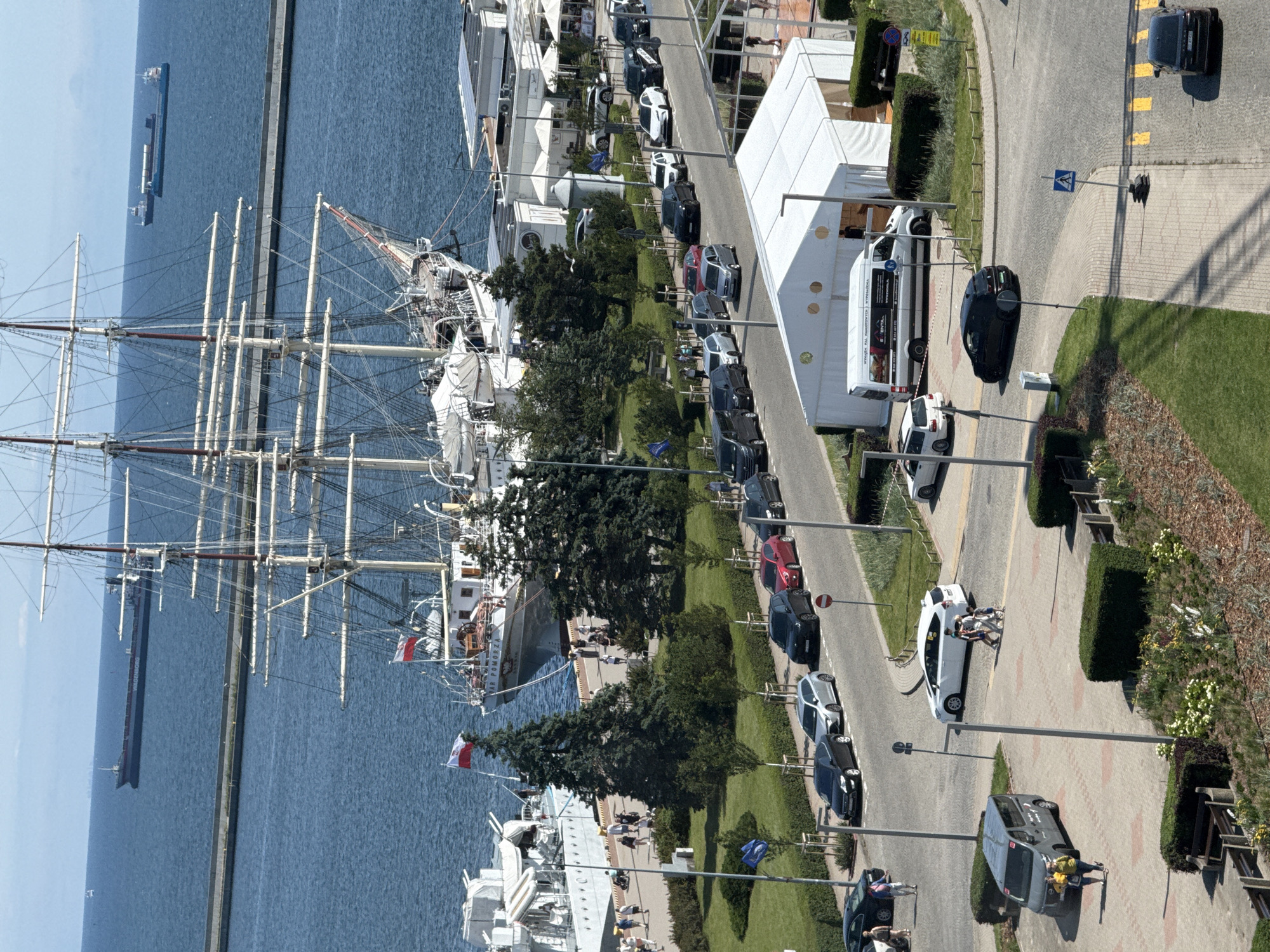 This is an aerial view of a harbor area in Gdynia, Poland, on a sunny day. The primary subject is a large, multi-masted sailing ship docked in the foreground, with its sails furled. To the left of the sailing ship, another vessel, likely a modern warship or patrol boat, is docked. The background shows the calm, blue water of the harbor, with a breakwater and several other ships, including a large cargo ship, visible in the distance.
Along the waterfront promenade, there are people walking and standing. Behind the promenade, there is a green grassy area with trees. A street runs parallel to the grassy area, lined with parked cars. Further down the street, there is a large white tent structure, possibly for an event, with a white van parked next to it. The street is busy with traffic. The scene is framed by a light blue sky with some scattered clouds.