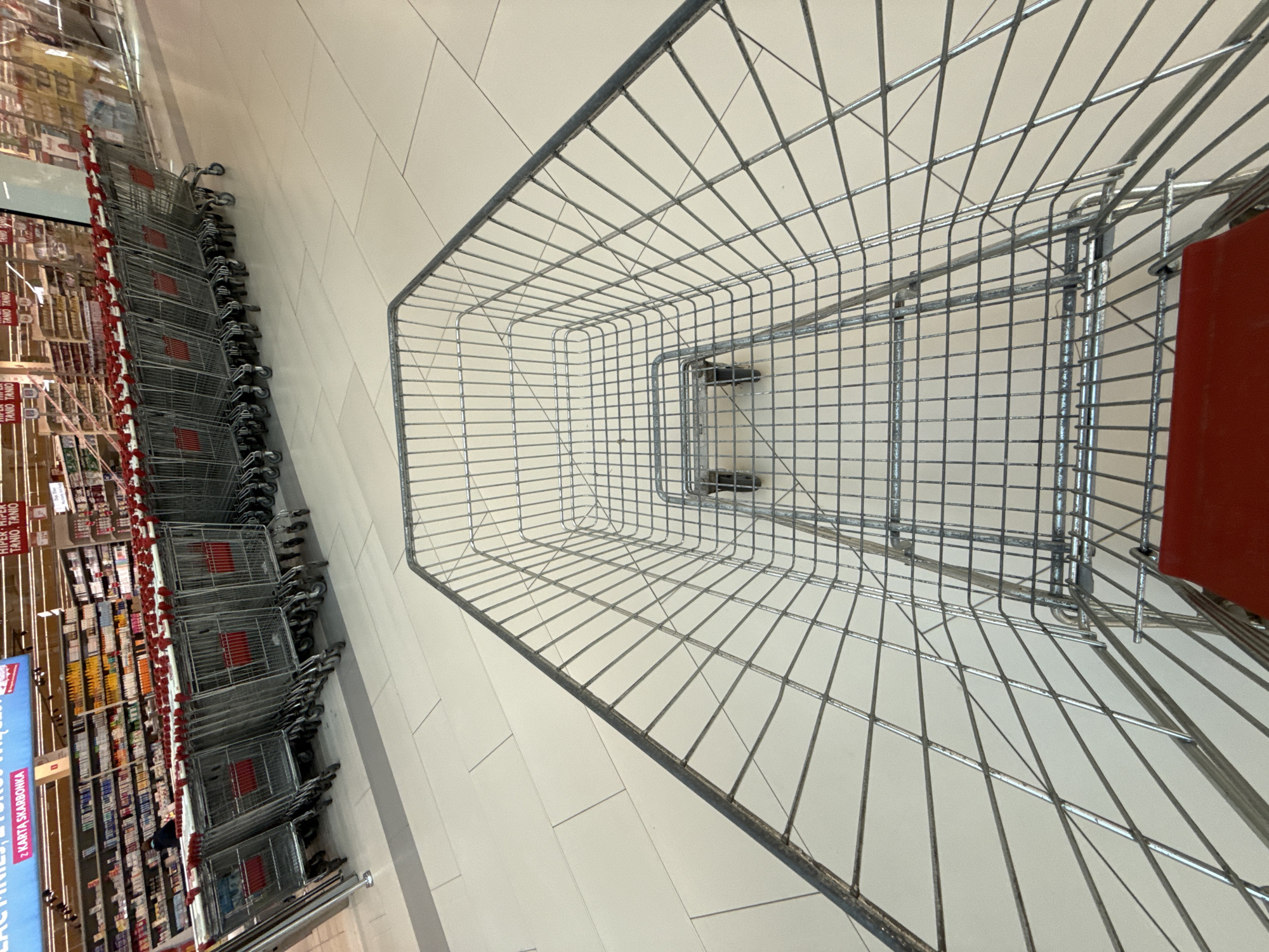 In a retail environment, a perspective shot is captured from within a shopping cart, looking forward. The floor is tiled with large, light-colored squares. In the background, a row of identical silver shopping carts, each with red plastic handles, are lined up. Behind the carts, shelves are filled with various products. Above the shelves, signage displays text in an unfamiliar language, including "HIPER HIPER TANIO TANIO" and "Z KARTĄ SKARBOK." A person can be partially seen near the front of the row of carts. The setting is identified as Bielany Wrocławskie, Poland.