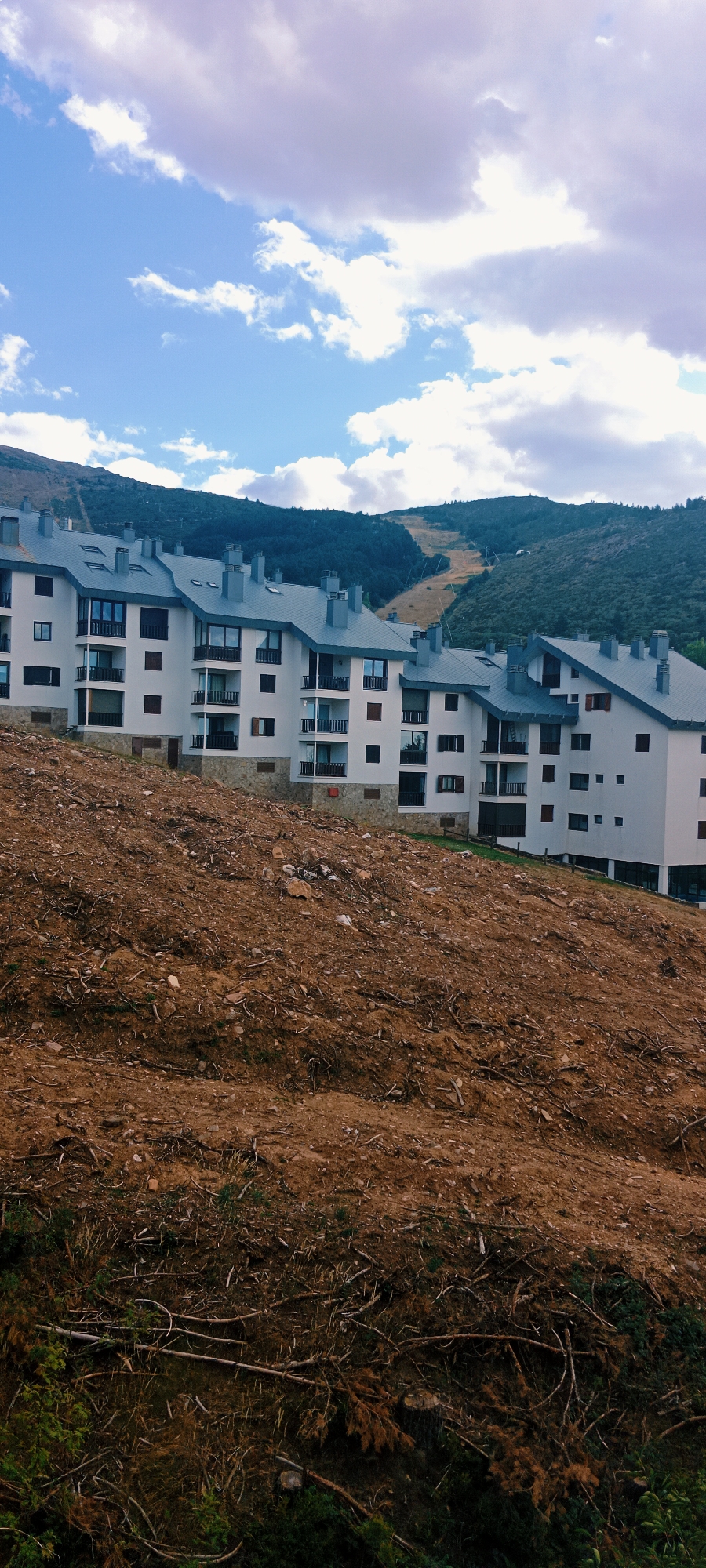 The image shows a residential complex located on a hillside in La Pinilla, Spain. The complex consists of multiple white buildings with dark grey roofs and numerous windows. The buildings are situated on a steep slope covered in reddish-brown soil, rocks, and scattered vegetation, indicative of recent landscaping or construction. In the background, a lush green mountain slopes upwards, with a visible ski slope or cleared path cutting through the trees. The sky above is a bright blue, dotted with fluffy white and grey clouds. There are no people visible in the image.