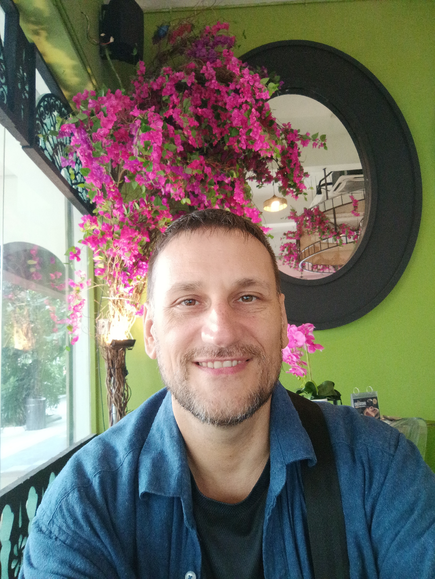 A man with short, dark hair and a beard smiles directly at the camera, his right side closer to the viewer. He wears a blue collared shirt over a dark t-shirt. Behind him, a large, round mirror with a black frame hangs on a lime green wall. A vibrant pink bougainvillea plant is positioned to the left of the mirror and extends upwards, with some of its branches reaching over the top edge of the frame. To the left of the bougainvillea is a window with a view of an outdoor area. The man appears to be indoors, likely in a restaurant or cafe, given the decor and a small table with items on it visible in the background to the right. The overall setting suggests a casual, possibly tropical, environment. Based on the architectural style of the railings visible to the left, the location appears to be in Kuala Lumpur, Malaysia, specifically within the historical district near Merdeka Square, known for its colonial-era buildings and decorative ironwork.