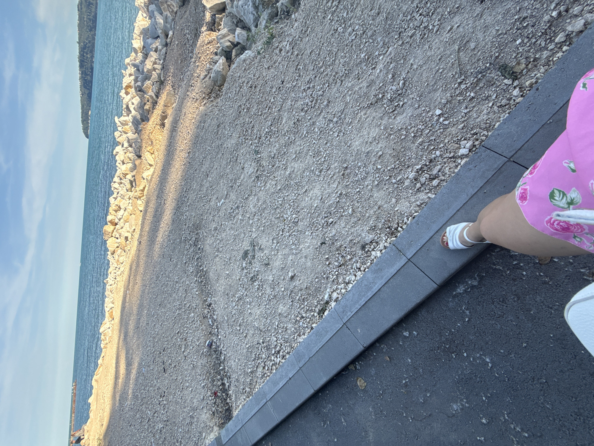 The image shows a person's legs and feet in the foreground, wearing white sandals and a bright pink floral dress. The person is sitting on an asphalt surface next to a concrete curb. Beyond the curb is a gravel path and a rocky shoreline leading to the sea. The sea is calm and blue, with a distant coastline visible. The sky is clear and blue with some scattered clouds. The overall setting appears to be a waterfront area, possibly a promenade or park, in the city of Varna, Bulgaria. The lighting suggests it is late afternoon or early evening.