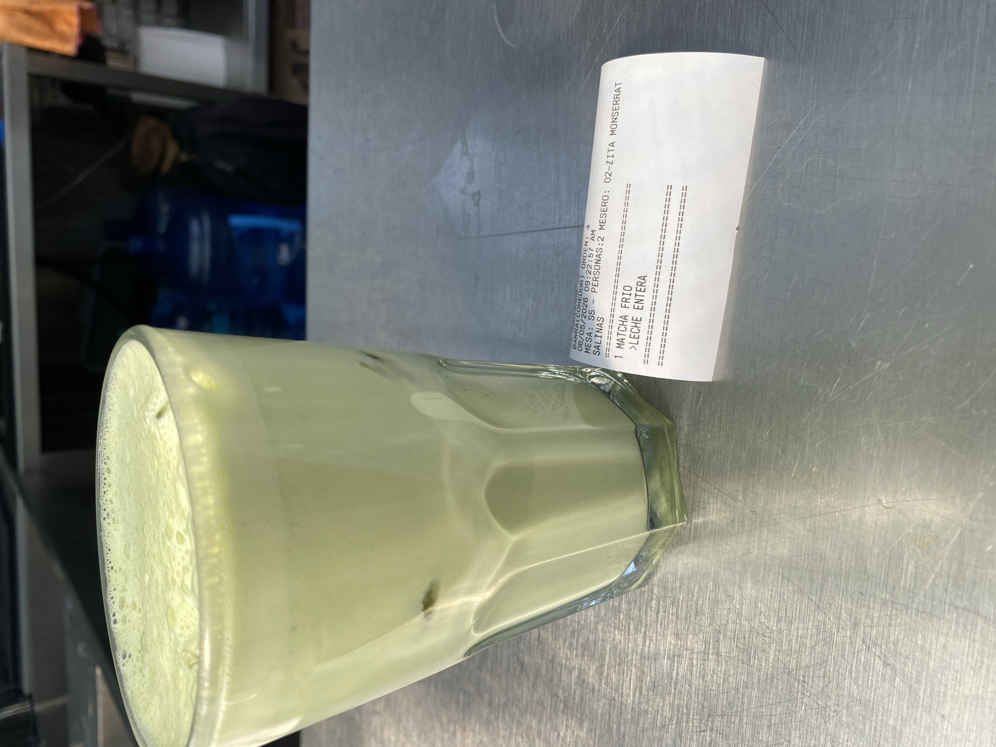 The image shows a glass of matcha latte and a receipt on a metallic surface. The receipt indicates the drink was purchased at "Salinas" and the restaurant is named "02-ZITA MONSERRAT". It was ordered on June 5, 2026, at 9:22:57 AM from "BARRA (COMEDOR)". The order was placed on Mesa S5 for 2 people. The drink is a "MATCHA FRIO" with "LECHE ENTERA" (whole milk). In the background are blue plastic water jugs.