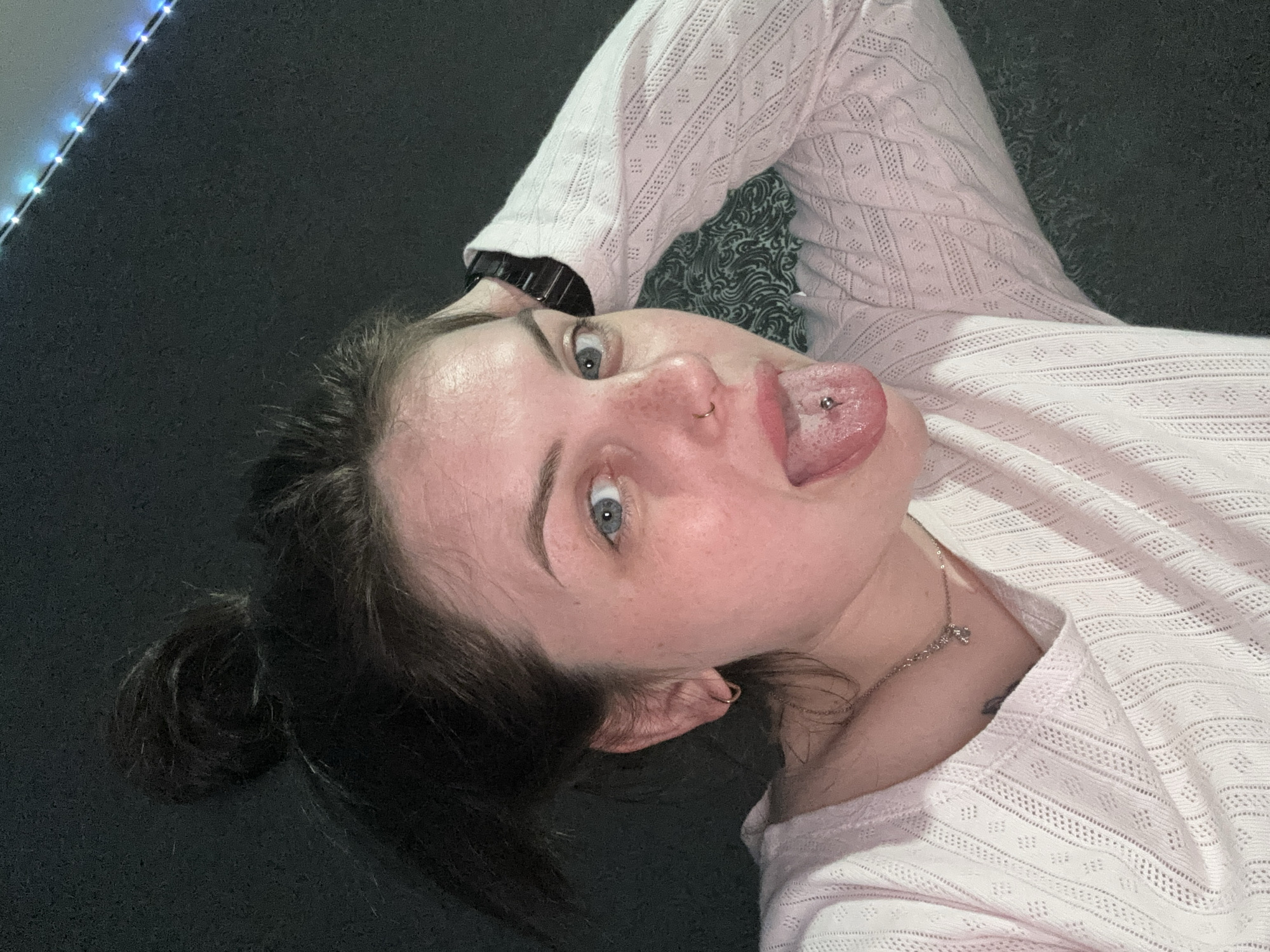 A fair-skinned woman with dark hair pulled back in a messy bun is lying on a dark surface. She is wearing a light pink, long-sleeved shirt with a decorative knit pattern. Her eyes are blue, and she has a nose ring and a tongue piercing. Her tongue is extended out of her mouth. She is also wearing a wristwatch and a silver necklace with a small charm. The background is dark, with a string of lights visible in the upper left corner. The location is not identifiable from the image.