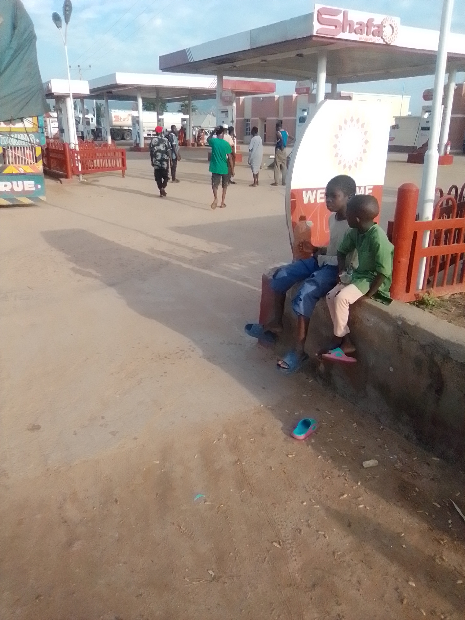 In Kwadon, Nigeria, two young boys sit on a concrete ledge in front of a gas station. The boy on the left, wearing a light grey t-shirt and blue pants, has a plastic bottle in his hand. The boy on the right, in a green t-shirt and light pink pants, looks to the left. Both are barefoot. Behind them, a sign displays "Shafa ENERGY" and a large circular emblem. The gas station has multiple pumps under a canopy, and several people are visible in the background, including a man in a red cap and black and white patterned shirt, and a man in a green t-shirt walking away from the camera. A truck with the word "RUE" visible on its side is parked to the left. The ground is a mixture of sand and dirt, with some debris scattered around, including a child's pink and blue shoe in the foreground. The sky is partly cloudy.
