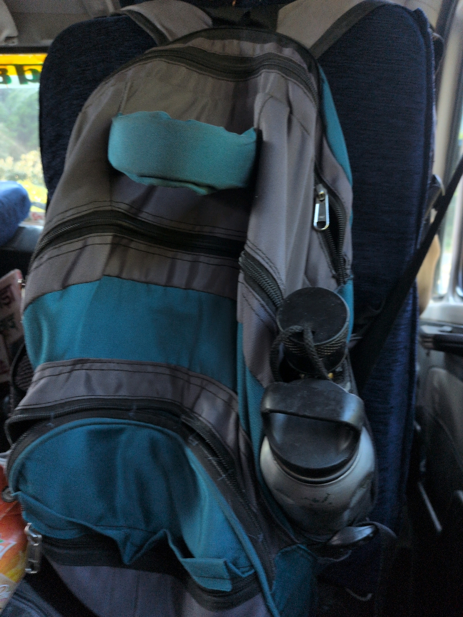 A backpack is hanging on a dark blue seat, likely inside a vehicle. The backpack is primarily gray and teal, with multiple zippered compartments and pockets. A bright teal fabric pouch is attached to the front of the backpack. Two black cylindrical objects, possibly water bottles or portable speakers, are attached to the side of the backpack with black cords. The background shows a blurred view of a window with some greenery and possibly signs, suggesting an outdoor scene beyond. The interior of the vehicle appears to be upholstered in dark fabric. The presence of a backpack and seating suggests transportation, and given the detail of a window with vegetation, it is likely that the image was taken while the vehicle was in motion. The location being Dharamshala, India, suggests a mountainous or hilly terrain.