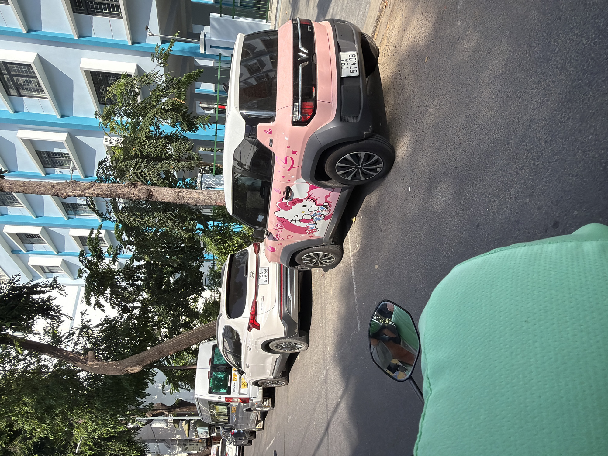 In Nha Trang, Vietnam, on a sunny day, a line of parked vehicles is captured on an asphalt road. A silver minivan with a license plate reading "31B 207.08" is in the foreground on the left. Next to it is a white Hyundai Santa Fe SUV, identified by the license plate "79A 428.98". To the right of the SUV is a pink VinFast VF 3 electric car adorned with a Hello Kitty decal and the license plate "79A 574.08". Behind these vehicles, a light blue building with white-trimmed windows is visible. Lush green trees line the street, casting shadows on the road and the cars. The perspective suggests the image was taken from a motorcycle, with a glimpse of the rider's green garment and a side mirror reflecting the scene. The background is dominated by the modern architecture of the blue building.