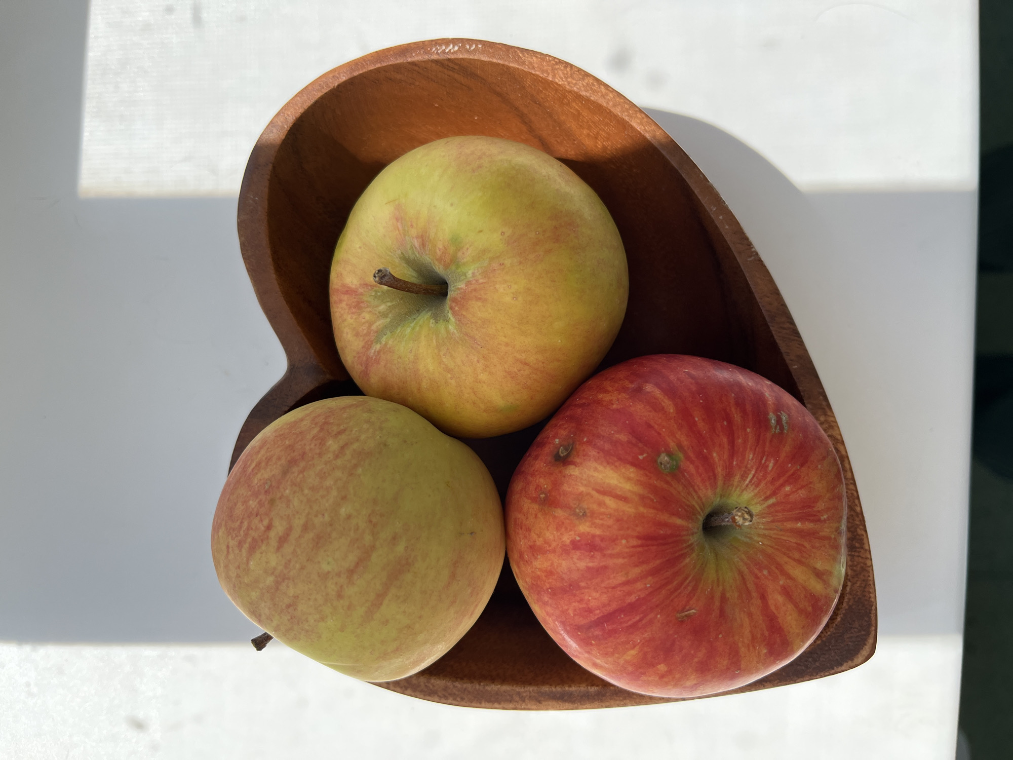 A close-up overhead shot displays three apples nestled within a dark wooden heart-shaped bowl on a white surface. The apples exhibit varying shades of red and yellow, with visible streaks and speckles. The lighting casts a shadow to the left of the bowl, suggesting illumination from the right. The scene is depicted with a shallow depth of field, focusing on the apples and the bowl. No specific location in Komsomolskoye, Ukraine, is identifiable from the image.