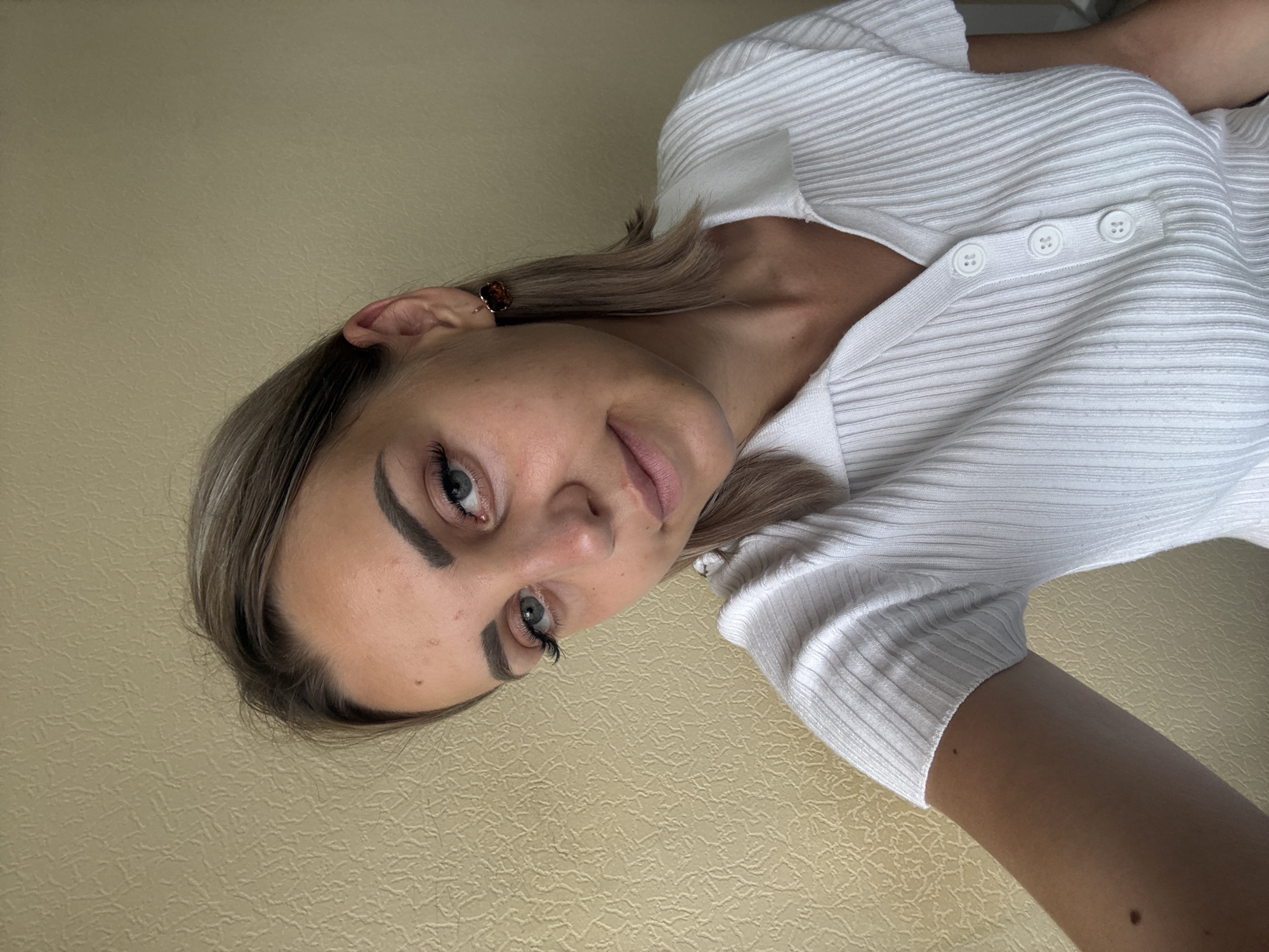A young Caucasian female with blonde hair and light blue eyes is pictured from the chest up, looking at the camera. She is wearing a white, ribbed polo shirt with a collar and three buttons. She has on a small, square earring with an amber-colored stone in her right ear. The background is a textured, light yellow wall. The image appears to be a selfie taken indoors. There is no visible indication of the location being Yuzhno-Sakhalinsk, Russia.