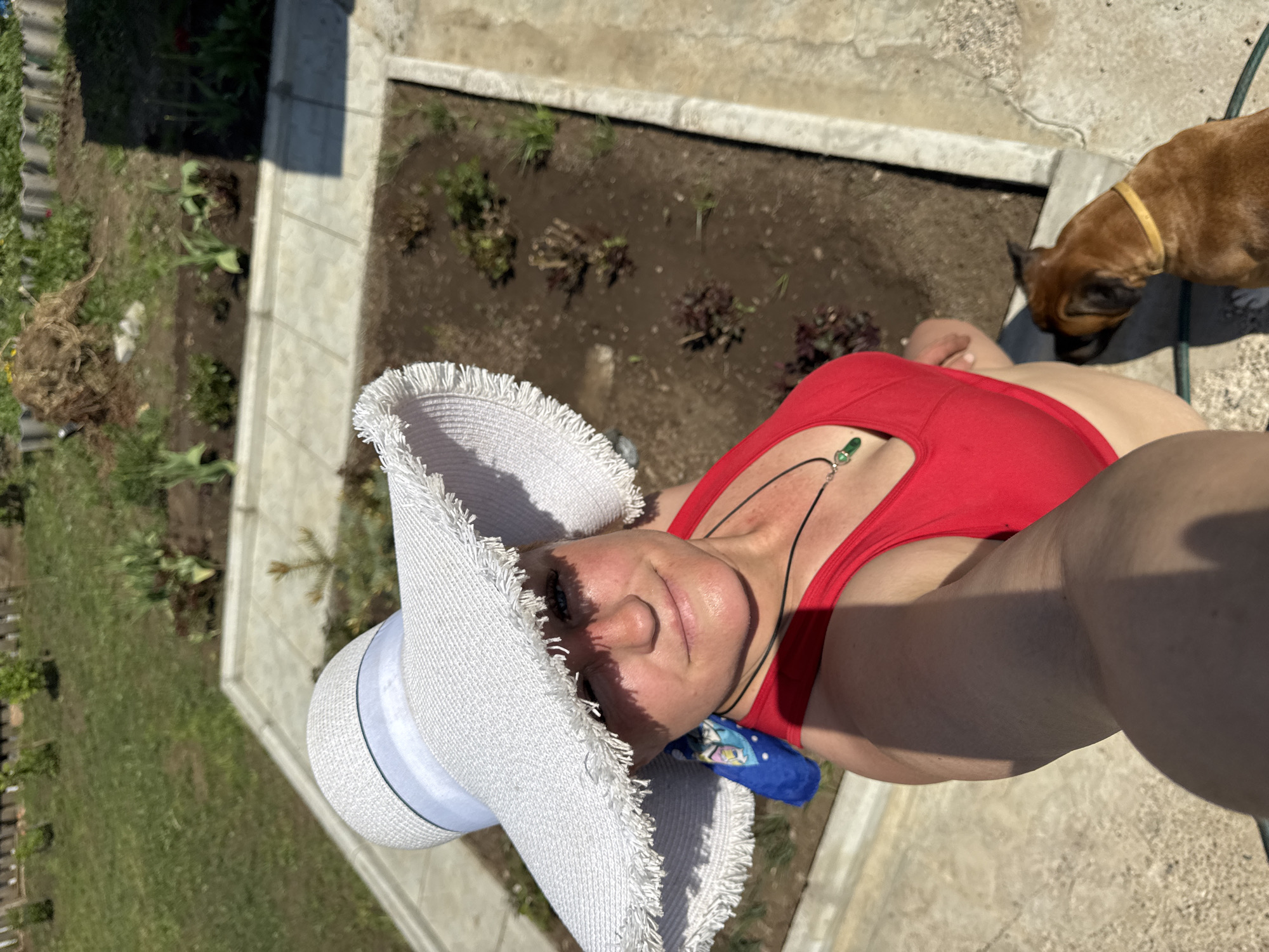 A woman wearing a wide-brimmed white hat and a red top is taking a selfie outdoors in Unknown, Unknown. The camera is tilted downward, capturing her face, part of her body, and some of the background. The woman has fair skin and is wearing a necklace with a green pendant. A blue scarf or bandana is tucked into the side of her top.
The background includes a garden bed with plants and soil. There is also grass and a fence visible in the distance. A brown dog with a yellow collar is partially visible in the upper right corner of the image. The lighting suggests it is daytime.