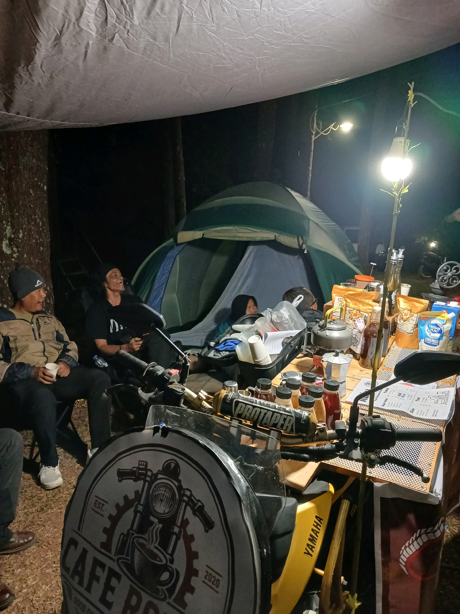 The image depicts a campsite at night in Berjo, Indonesia, possibly a roadside cafe setup. Several people are present. In the foreground, a motorcycle is parked next to a table laden with coffee-making supplies, bottles of drinks, and packaged goods. A tarp-covered structure provides shelter overhead, and a tent is visible in the background. The lighting is provided by artificial lamps, creating a warm and inviting atmosphere. Two men are seated in chairs, appearing to be engaged in conversation. One man is holding a cup, while the other is smiling. A woman is seated inside the tent, partially visible. The environment is rustic, with trees and natural ground cover surrounding the campsite. The overall scene suggests a casual gathering or a small business operating outdoors in a natural setting.