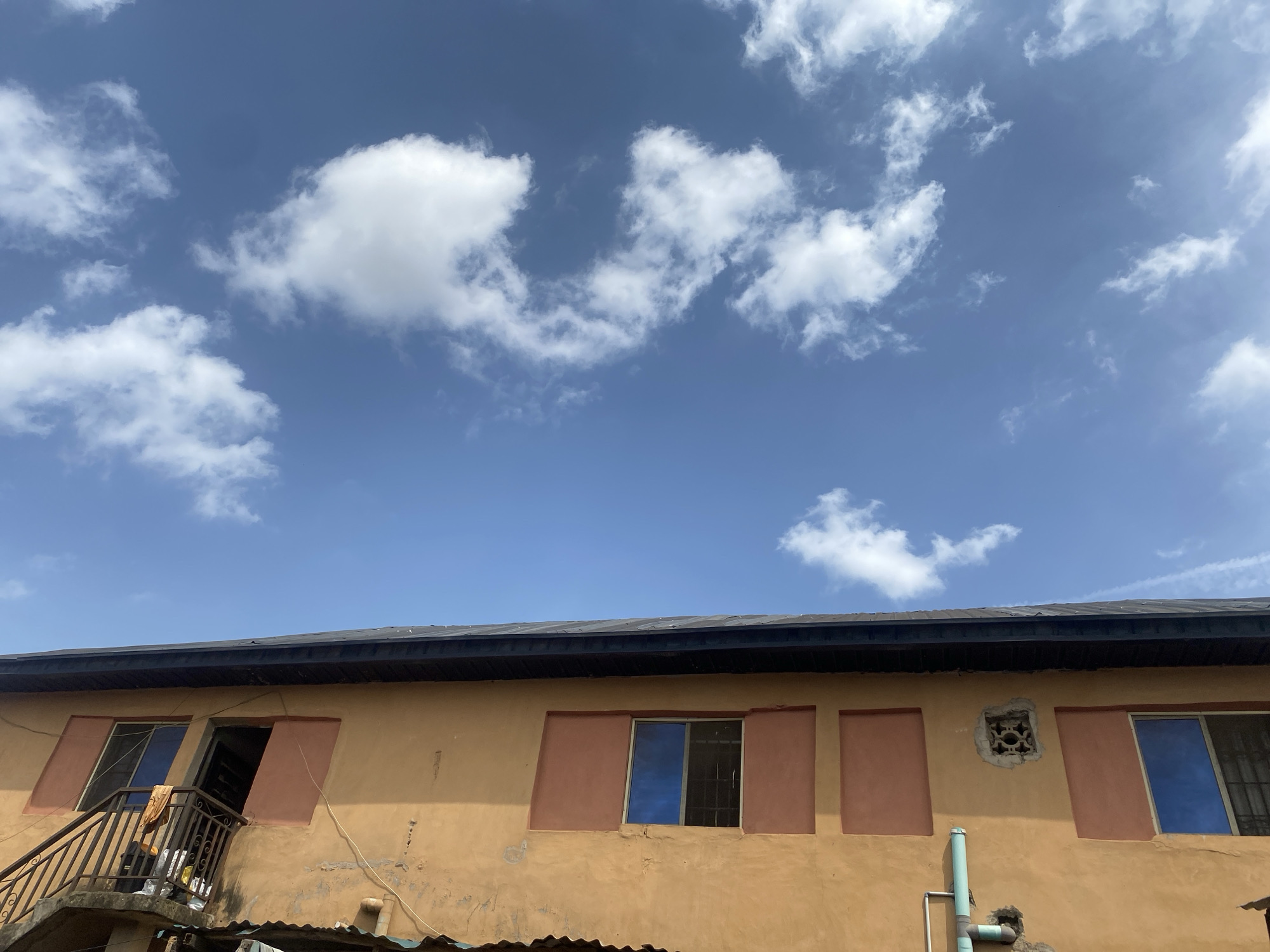 This image presents an exterior view of a building in Lagos, Nigeria, under a bright, sunny sky. The top half of the frame is dominated by a clear blue sky dotted with scattered white cumulus clouds.
The lower half of the image focuses on the upper portion of a multi-story building. Its facade is painted a warm, muted orange or yellow, with several rectangular sections around openings highlighted in a contrasting reddish-brown. On the left, a dark metal balustrade for a staircase or balcony leads to an open doorway. On the balcony, draped clothing and various items are visible. Adjacent to this, a window features a blue-tinted covering. Further along the wall, another window also has a blue tint, with visible metal bars behind it. A closed-off section with a reddish-brown panel is next, followed by a small, square opening or vent with a decorative metal grille set into the wall. Below this, light blue/green pipes run down the side of the building. The roof is dark-colored, appearing to be made of metal sheets, and glimpses of corrugated metal roofing are visible at the very bottom of the frame, possibly from a lower structure.
The scene depicts a clear, bright day with strong sunlight. No people are visible, and there is no active event taking place, presenting a static architectural scene in an urban or semi-urban environment. No text is visible within the image.