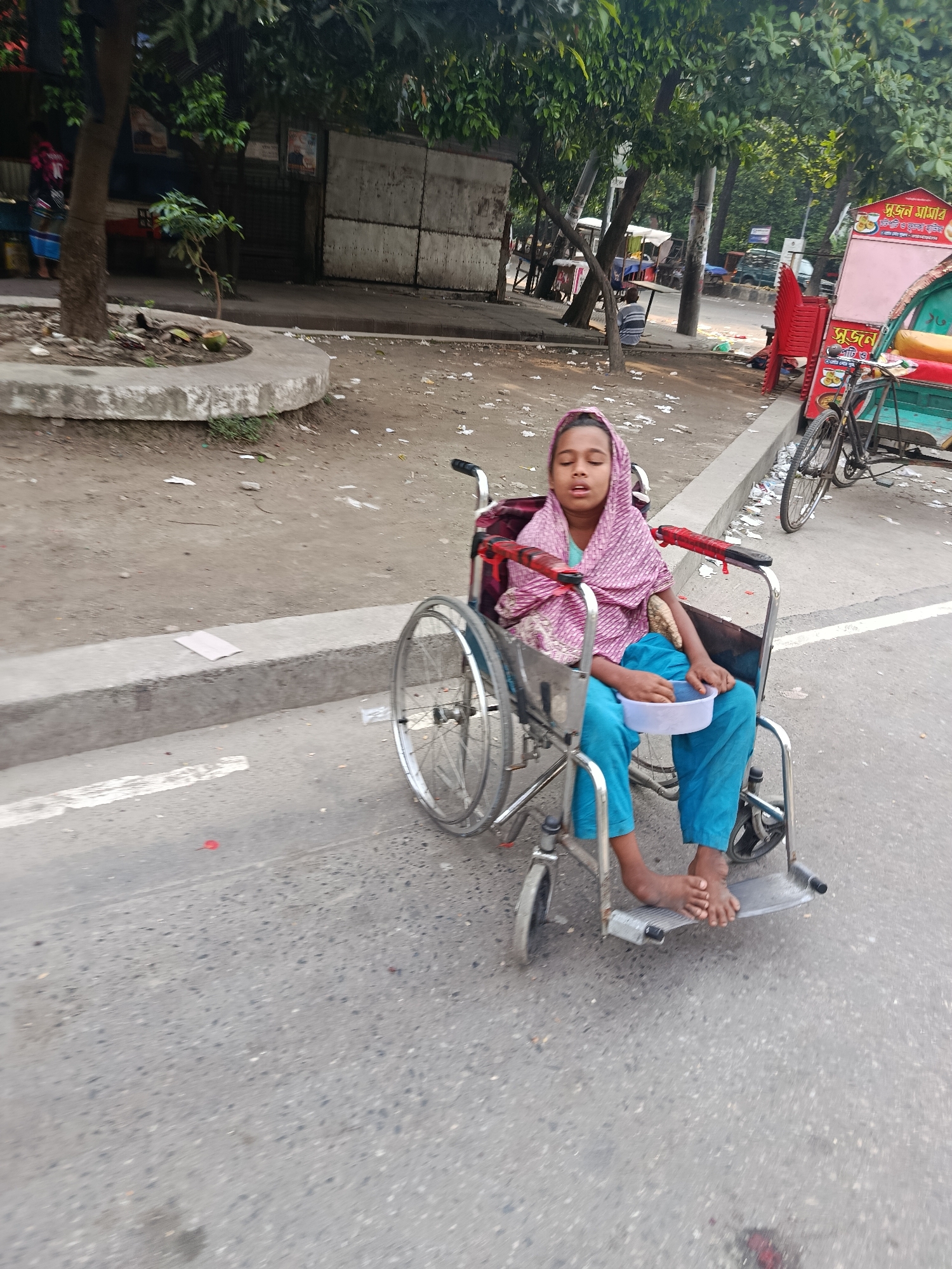 A young girl sits in a wheelchair on a city street in Dhaka, Bangladesh. She wears a light pink headscarf and blue clothing. She holds a white bowl in her lap. The wheelchair is on the pavement of a road. Trees and buildings are visible in the background. A three-wheeled vehicle and bicycle are parked nearby. The ground is littered with debris.