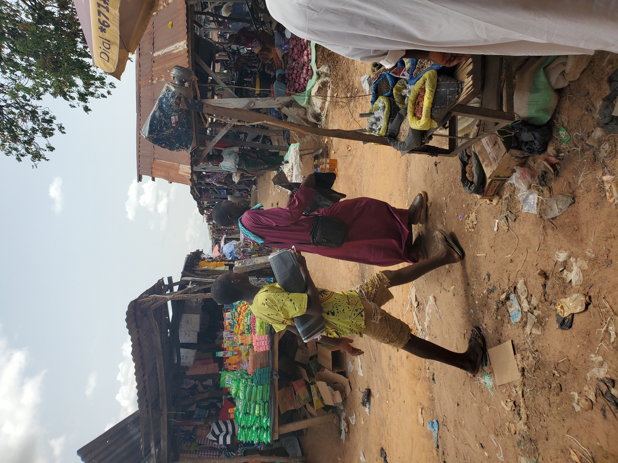 The image captures a street scene in Mararaba, Nigeria. The environment is a bustling marketplace with various stalls displaying goods. People are walking along the sandy or dirt path between the stalls. A young boy in a yellow shirt and tan shorts carries a cylindrical object while walking alongside an adult in a maroon robe. The stalls are made of wood and corrugated metal, with a variety of products on display. Trash and debris are scattered on the ground.