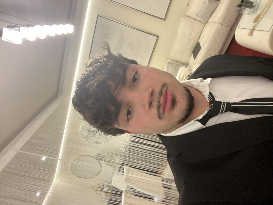 A young man in a black suit with a white shirt and black-and-white tie poses for a selfie. He has dark curly hair and a light beard. The background features a modern interior with a white couch, a wall decoration with hanging strands and circular mirrors, and a contemporary chandelier. The scene suggests a luxurious or upscale setting.