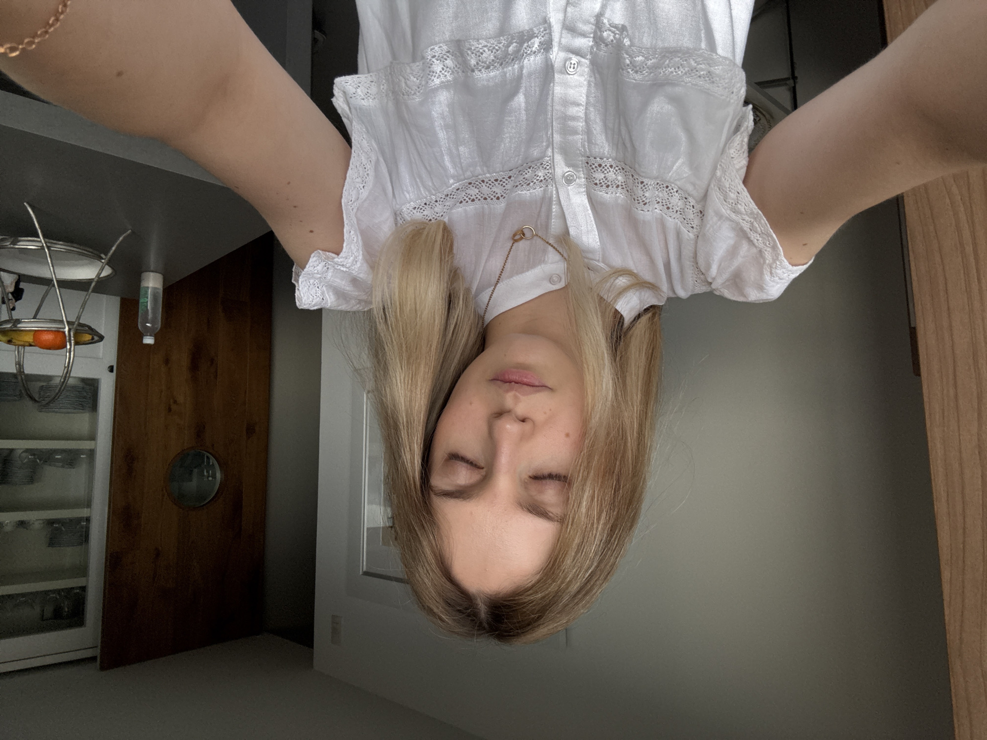 A woman is seen upside-down, presumably taking a selfie. She has long blond hair and is wearing a white blouse with lace trim and a gold necklace. Her eyes are closed. Her arms are outstretched to either side.
The background includes a gray countertop, a cabinet with glassware and fruit, a wooden sliding door with a round window, a gray wall, and a wooden door frame.
There is no indication of the location being Mexico City, Mexico.