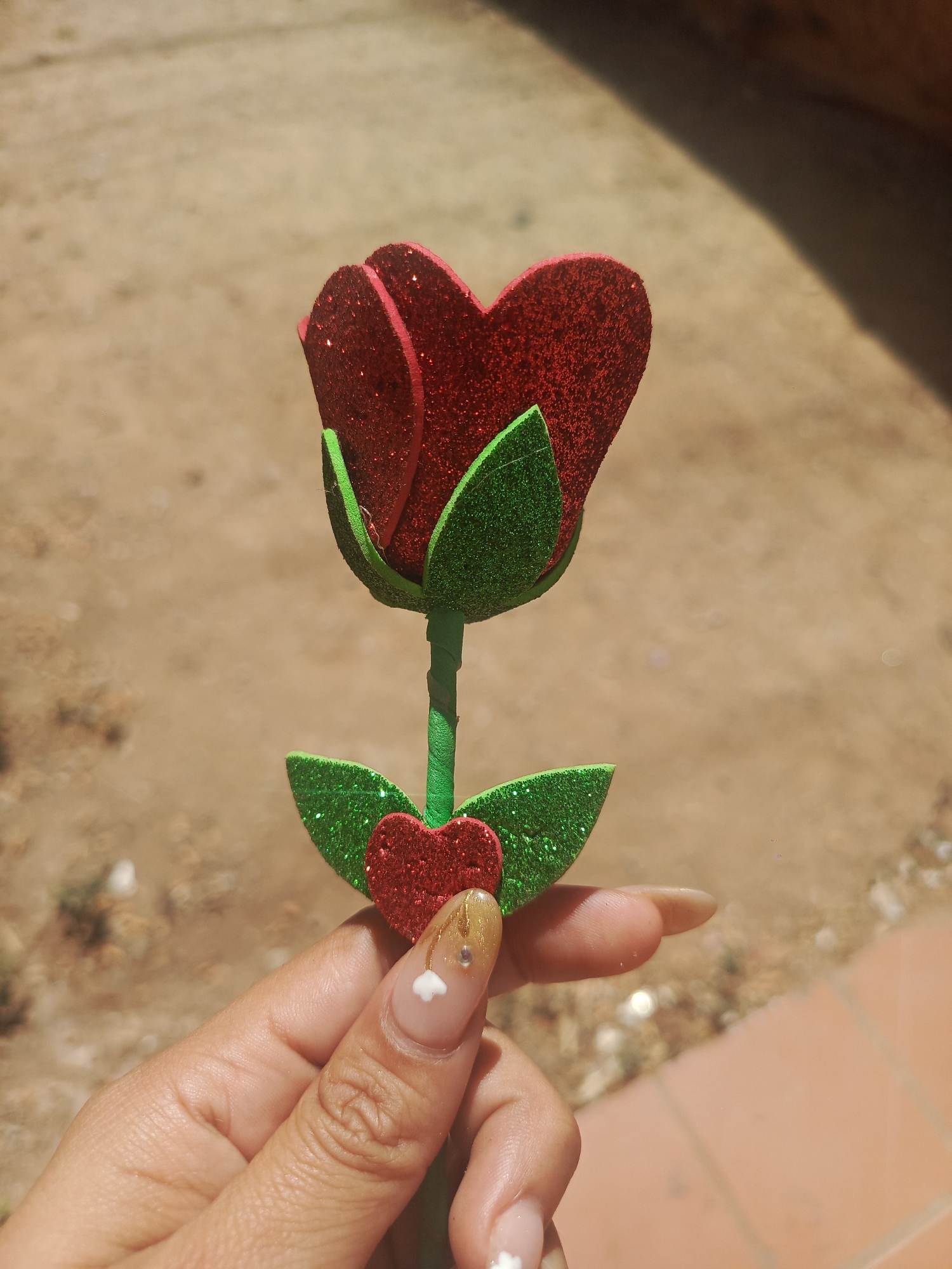 A hand is holding a decorative rose. The rose is constructed from red and green glitter-covered foam material, shaped into heart-like petals and leaves. The background consists of a blurred mix of a dirt-like surface and tiled floor. The location is likely outdoors, and based on the instructions, specifically in Juan Griego, Venezuela.