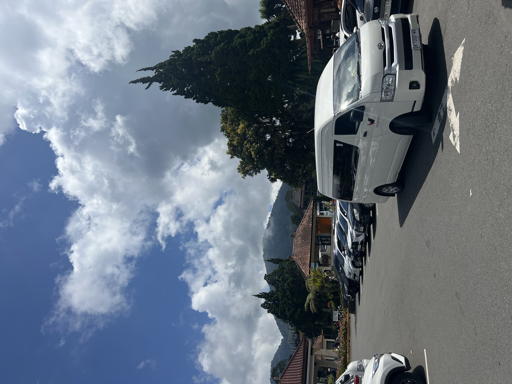 The image captures a street scene in Baturiti, Indonesia. A row of parked cars and vans lines the side of the road, with a white van prominently displayed at the forefront. In the background, tiled roofs of buildings are visible, interspersed with lush green trees. A mountain range rises in the distance, partially obscured by clouds. The sky overhead is a mix of blue patches and puffy white clouds. The scene is bathed in daylight, suggesting a daytime setting.