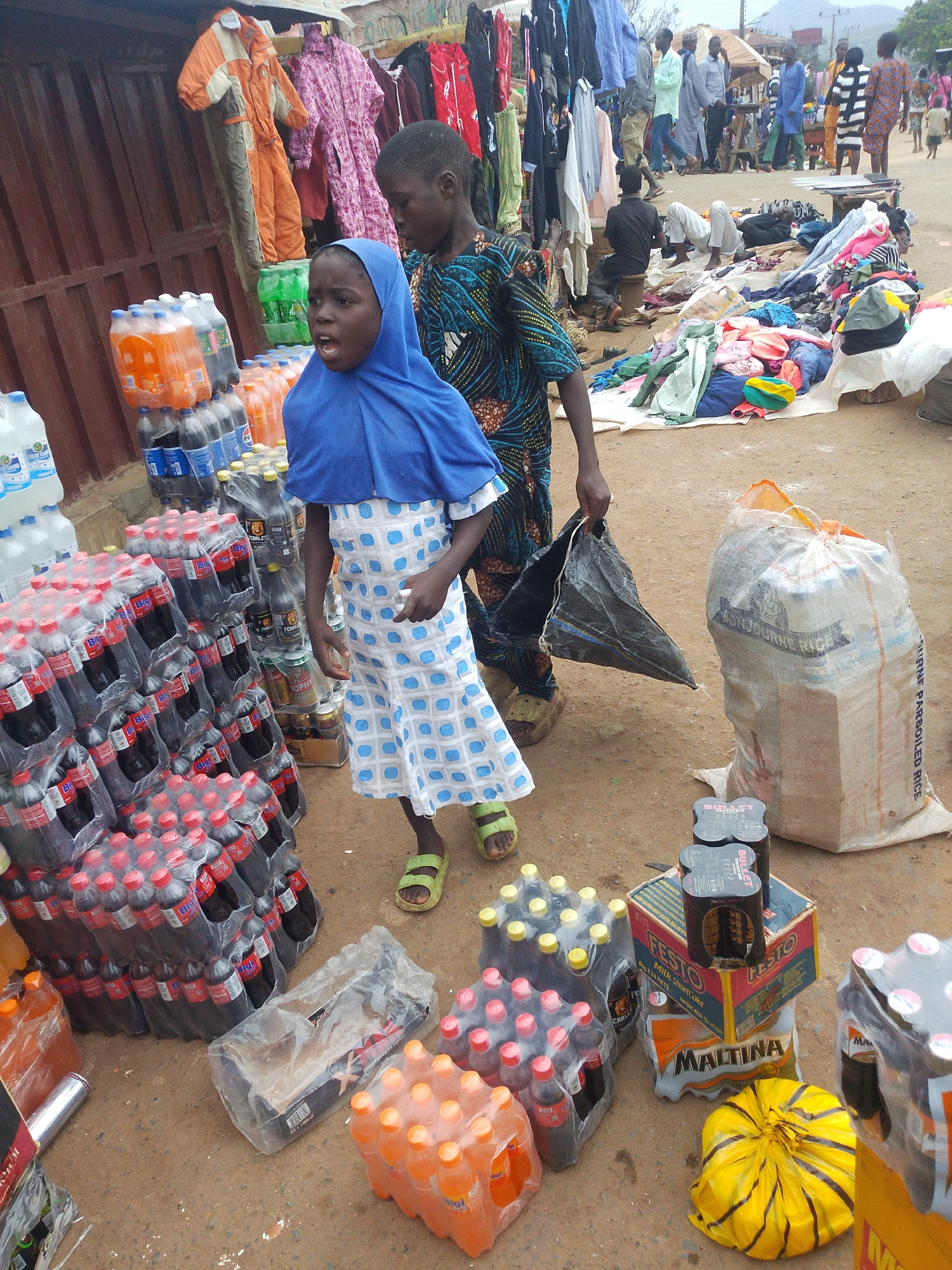 In Komu, Nigeria, two young individuals are at the forefront of the image, surrounded by numerous stacked bottles of drinks and sacks. A young girl, wearing a blue hijab and a white dress with blue polka dots, stands in the foreground, her mouth open in an expression of surprise or excitement. Beside her, a young boy in a patterned shirt appears to be walking away, carrying a black plastic bag.
The ground is a dusty, reddish-brown dirt, indicative of a market setting. Behind the children, a stall displays various clothing items, including an orange and grey jumpsuit and brightly colored garments. The scene is filled with merchandise, predominantly beverages in plastic bottles and in large sacks. Several brands of drinks are visible, including Coca-Cola and Maltina. A large sack, labeled with what appears to be rice, is prominently placed to the right of the children. In the background, more people and stalls are visible, suggesting a bustling marketplace. The overall atmosphere appears lively and commercial.