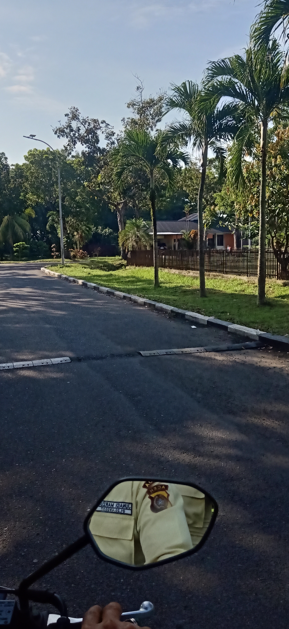 The image depicts a street scene in Tambontunong, Indonesia, on a clear day with a bright sky. A paved road dominates the foreground, with a white and grey curb lining the right side. Two speed bumps are visible on the road. The left side of the road features a grassy verge, a sidewalk, and a line of trees. In the background, a house with a dark roof and wooden fencing is visible behind a row of palm trees and other lush vegetation. The overall impression is a well-maintained suburban or semi-rural environment.