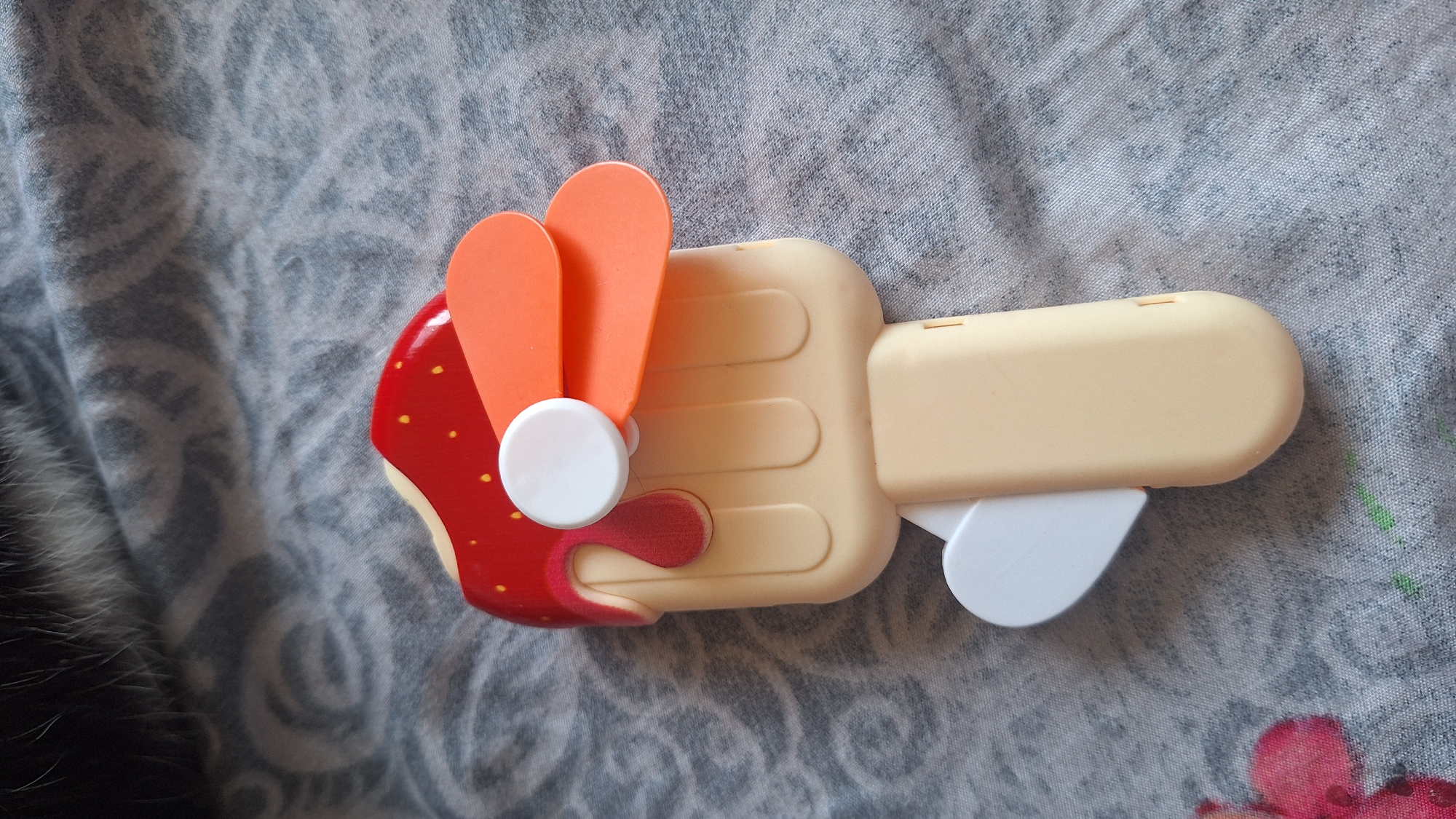 A close-up, high-angle shot shows a toy popsicle-shaped fan resting on a gray patterned fabric. The fan's "ice cream" top is red with yellow speckles, resembling a strawberry. Two orange fan blades are attached to a white circular component on the top. The handle of the fan is a pale yellow, with a white heart-shaped element on one side near the bottom. The fabric appears to be a bedsheet or tablecloth with a swirling, abstract pattern. No specific location in Samara, Russia is identifiable from the image.