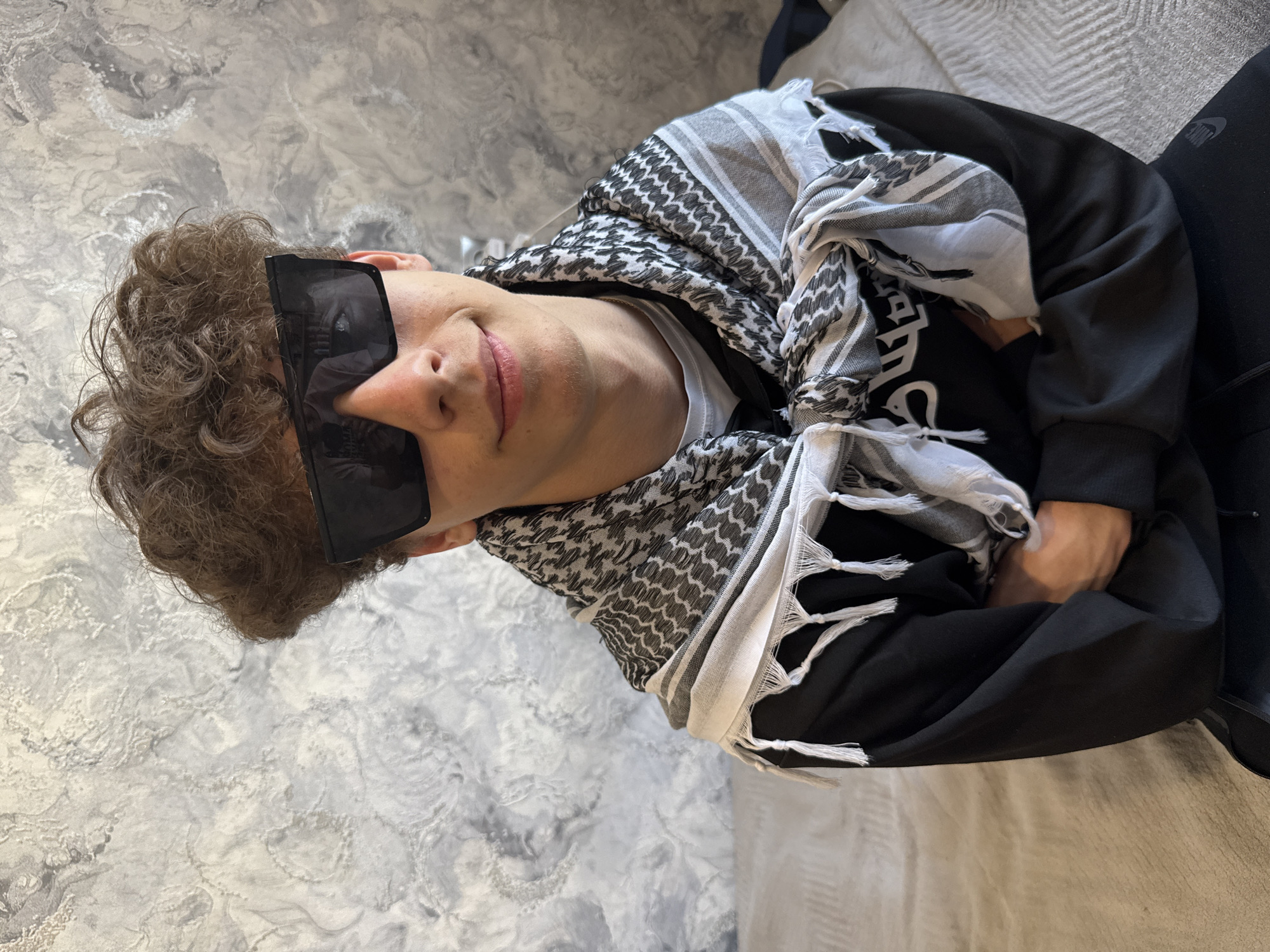 The image shows a person lying on a bed, wearing sunglasses and a black and white checkered keffiyeh scarf. The person is wearing a black hoodie with white writing visible. The background features a textured wall, possibly plaster or concrete. A portion of the bed is visible with light grey bedding. The location cannot be determined from the image.