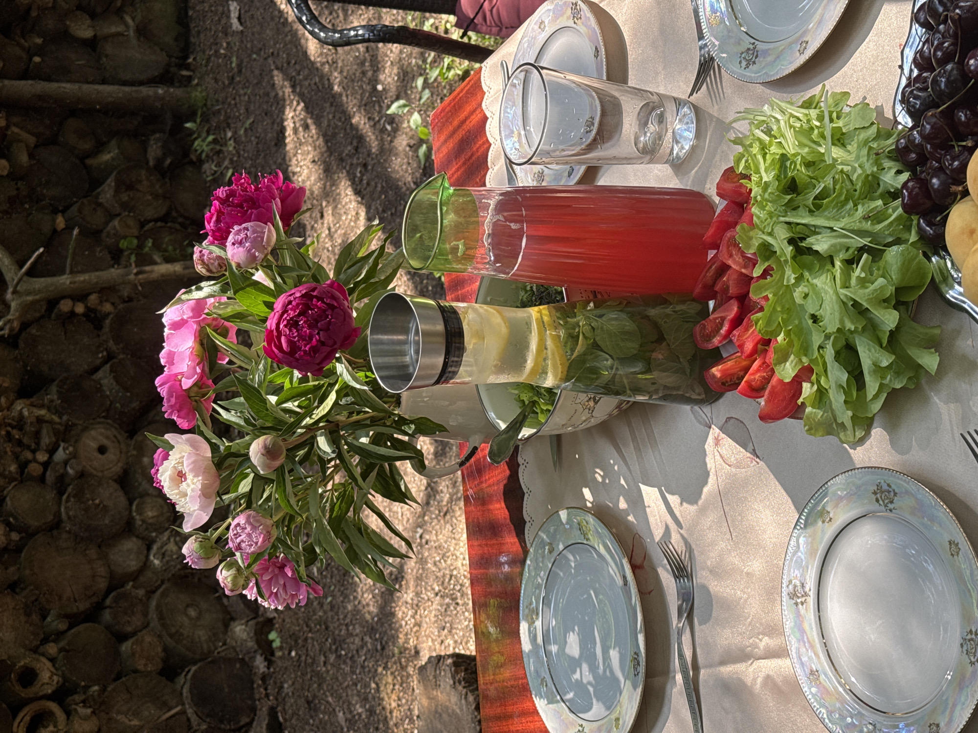 A table is set for an outdoor meal in Козино, Russia. A bouquet of pink peonies sits in a white pitcher. Nearby, a glass carafe holds lemon and mint infused water, and a green pitcher contains red juice. Plates, forks, a salad, chopped tomatoes, and a bowl of cherries are arranged on a tablecloth. The background features a stack of logs. The scene is bathed in sunlight, casting shadows.