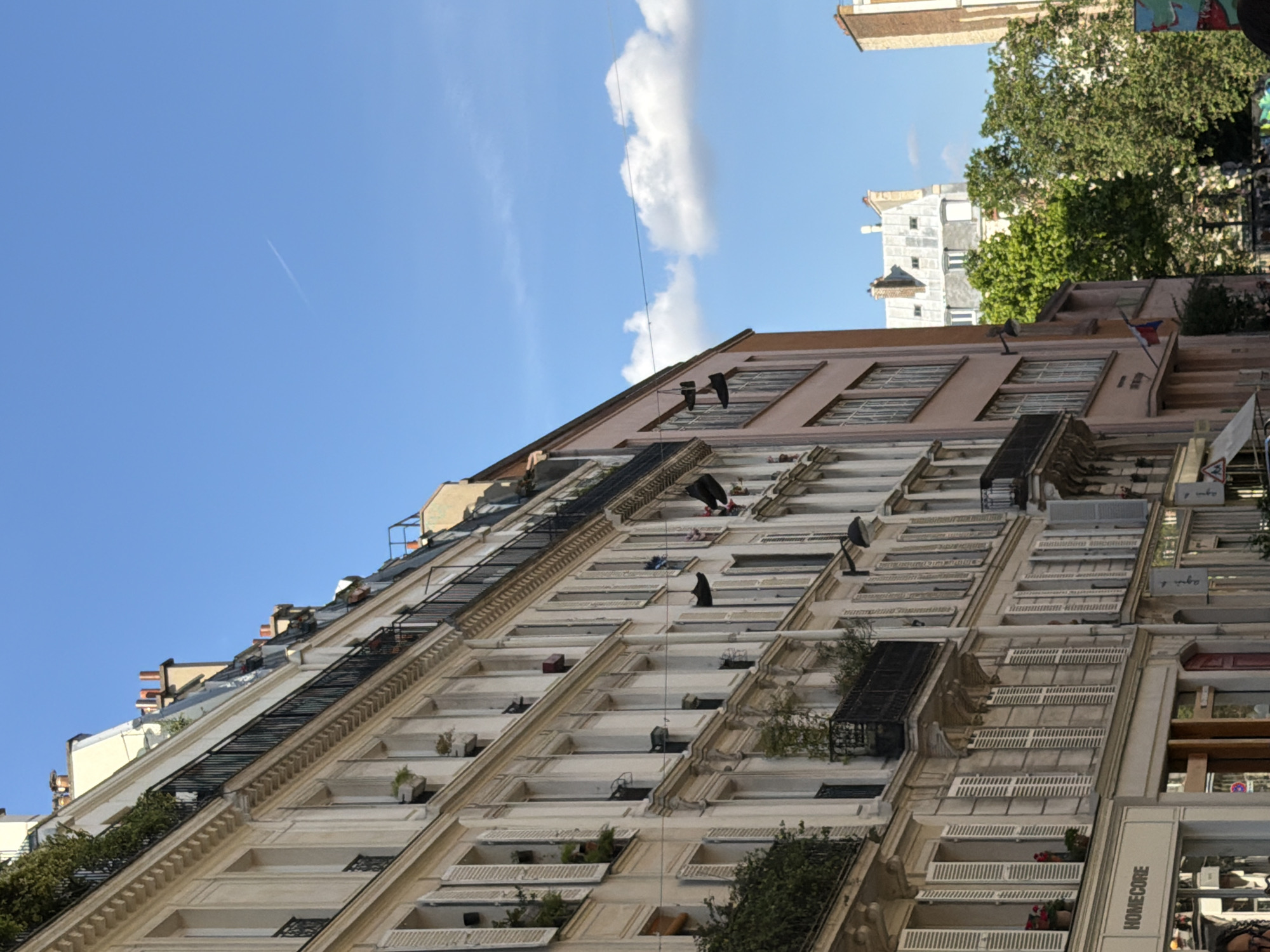 A low-angle shot of a tall, light-colored building in Paris, France, featuring multiple rows of windows and balconies with ornate detailing. The sky is blue with a thin contrail and a white cloud. Some balconies have plants. On the building's right, there is a tree and the top of a commercial storefront. Some laundry hanging from the top. The building has a fire escape.